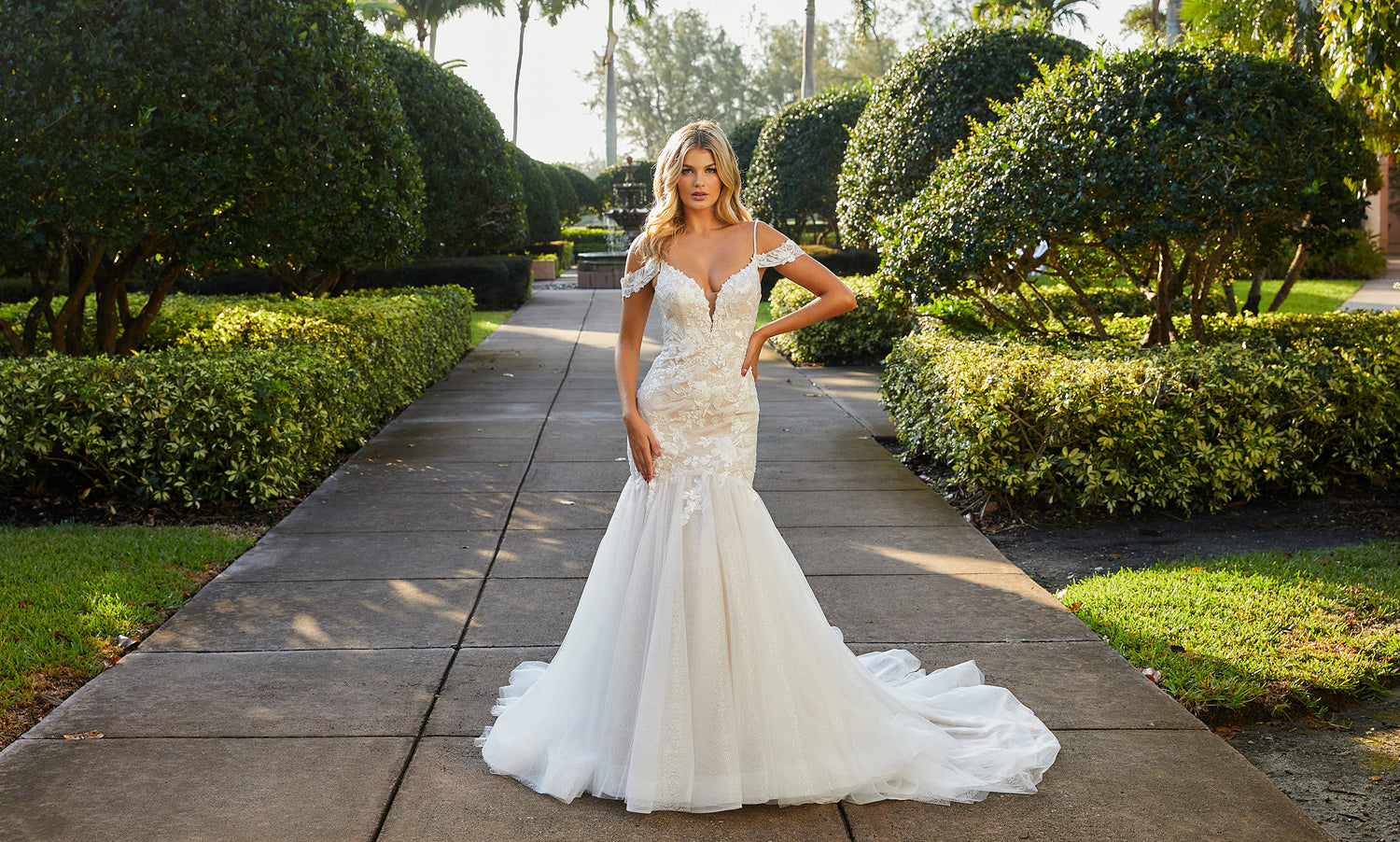 Bride in an off-the-shoulder lace mermaid gown on a sunlit hedge-lined garden walkway