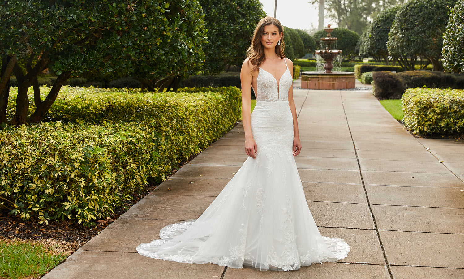 Bride in a V-neck lace mermaid gown with thin straps standing on a tree-lined garden walkway