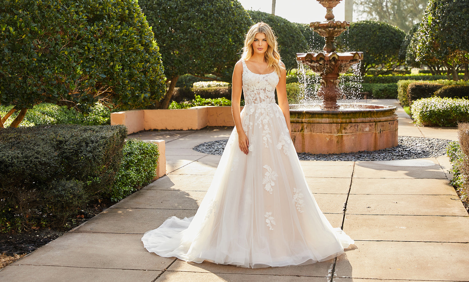 Bride in a scoop-neck lace and tulle A-line gown standing on a garden path by a fountain