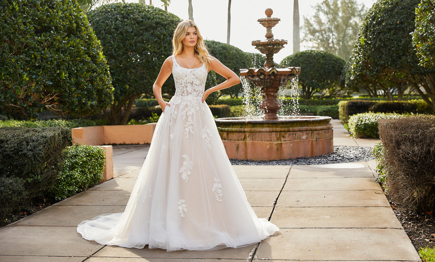 Bride with hands on hips in a scoop-neck lace and tulle A-line gown on a garden path