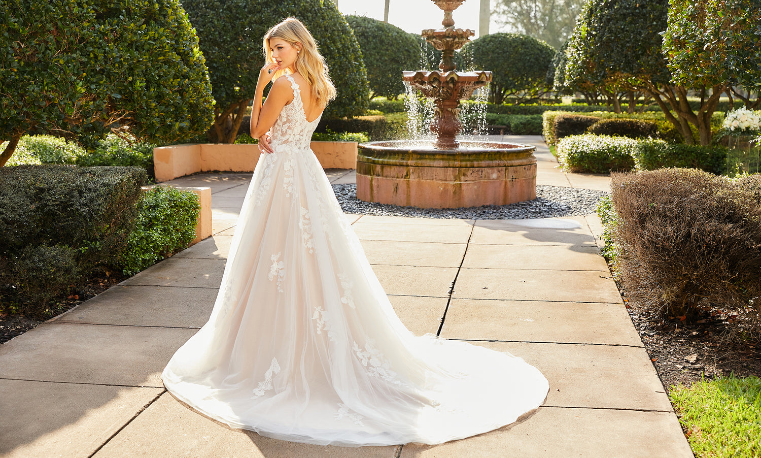 Bride seen from behind glancing over her shoulder in a blush lace A-line gown near a garden fountain