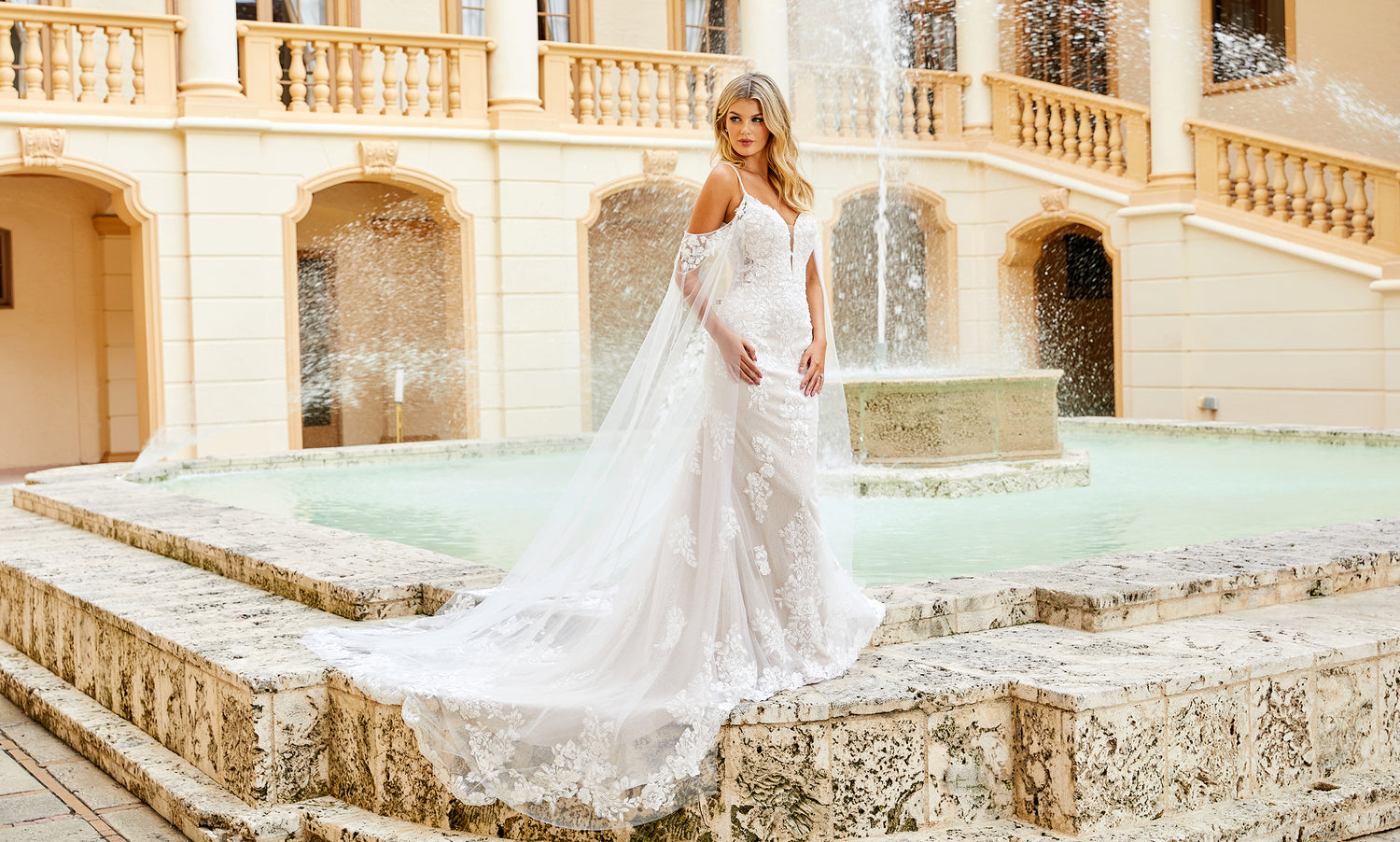 Bride in an off-shoulder lace mermaid gown standing at the edge of a stone fountain pool