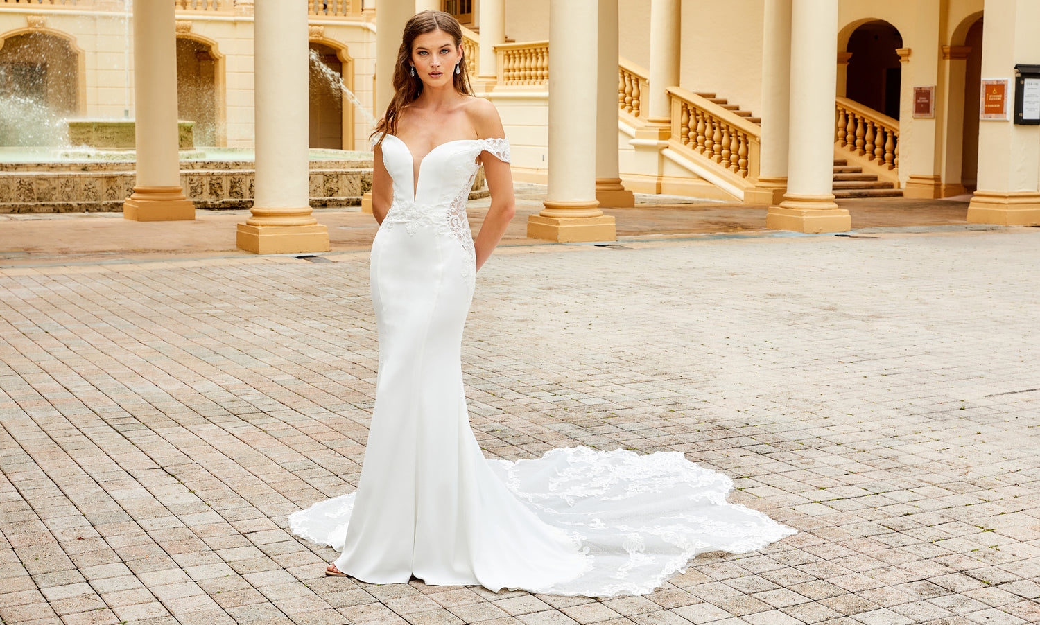 Brunette bride in an off-shoulder white crepe mermaid gown with a lace train on a brick courtyard