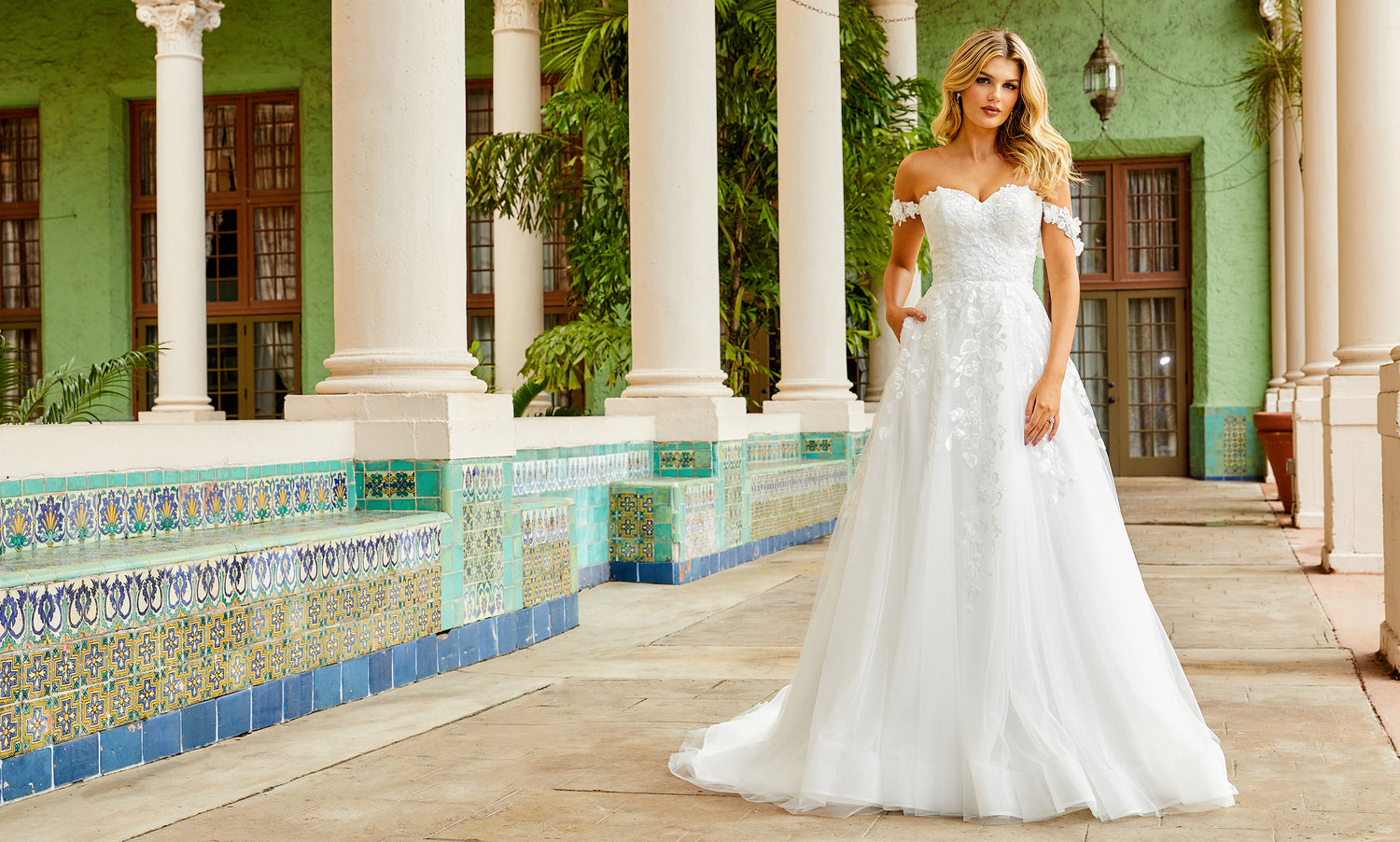Bride in an off-shoulder white lace ball gown with pockets, hands tucked in, beside a green colonnade