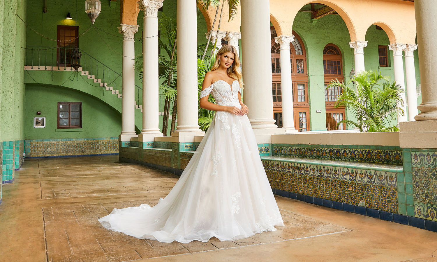 Bride in an off-shoulder white lace ball gown standing in a green tiled arcade with palms
