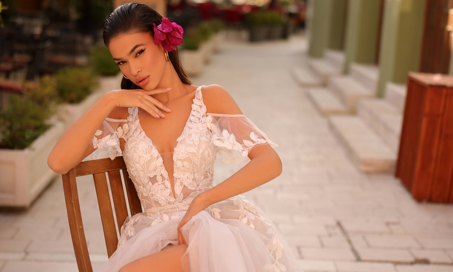 Dark-haired model seated on a wooden chair in a plunging floral-appliqué gown, pink flower in her hair