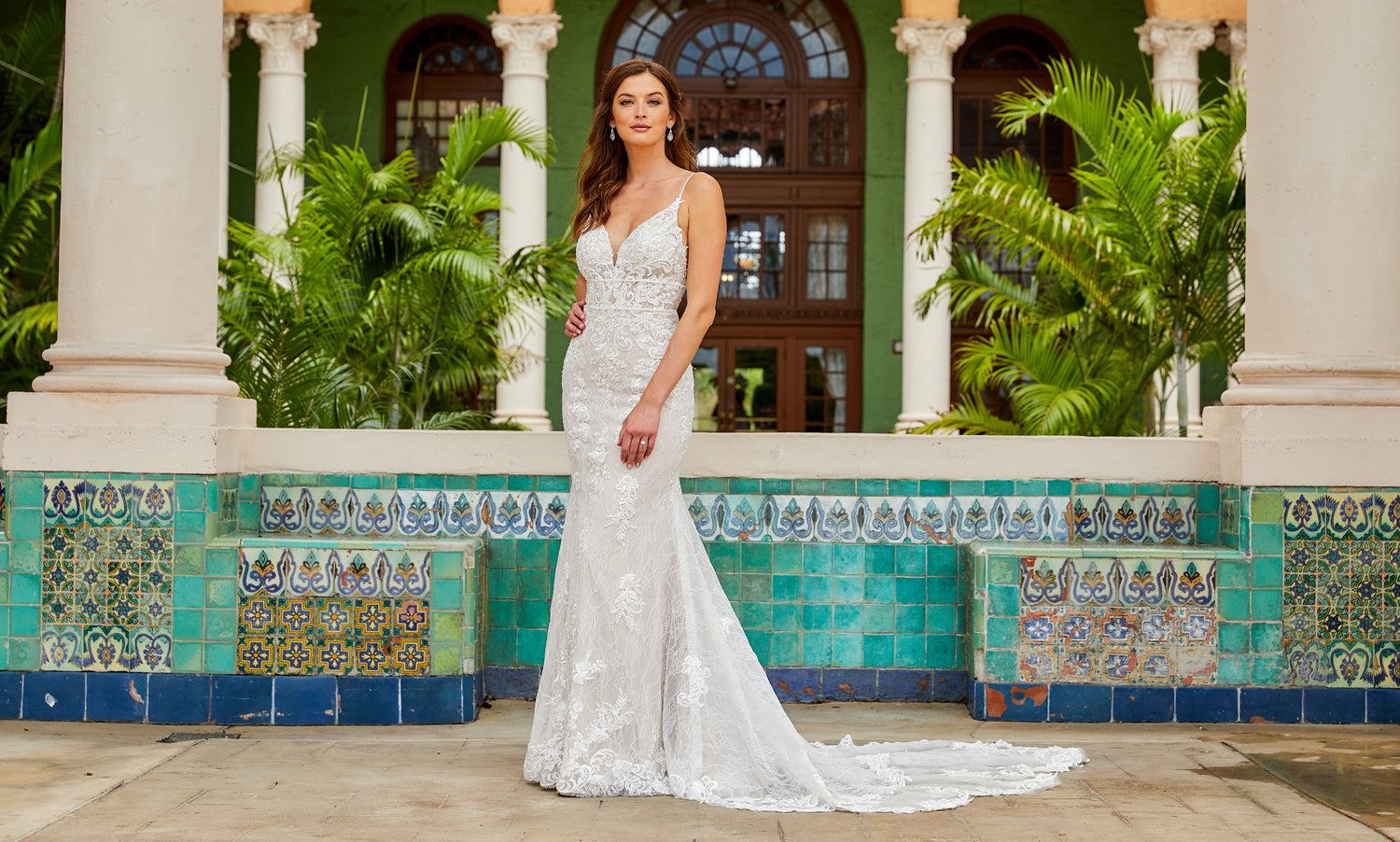 Brunette bride in a white lace mermaid gown standing before a turquoise tiled wall and palms