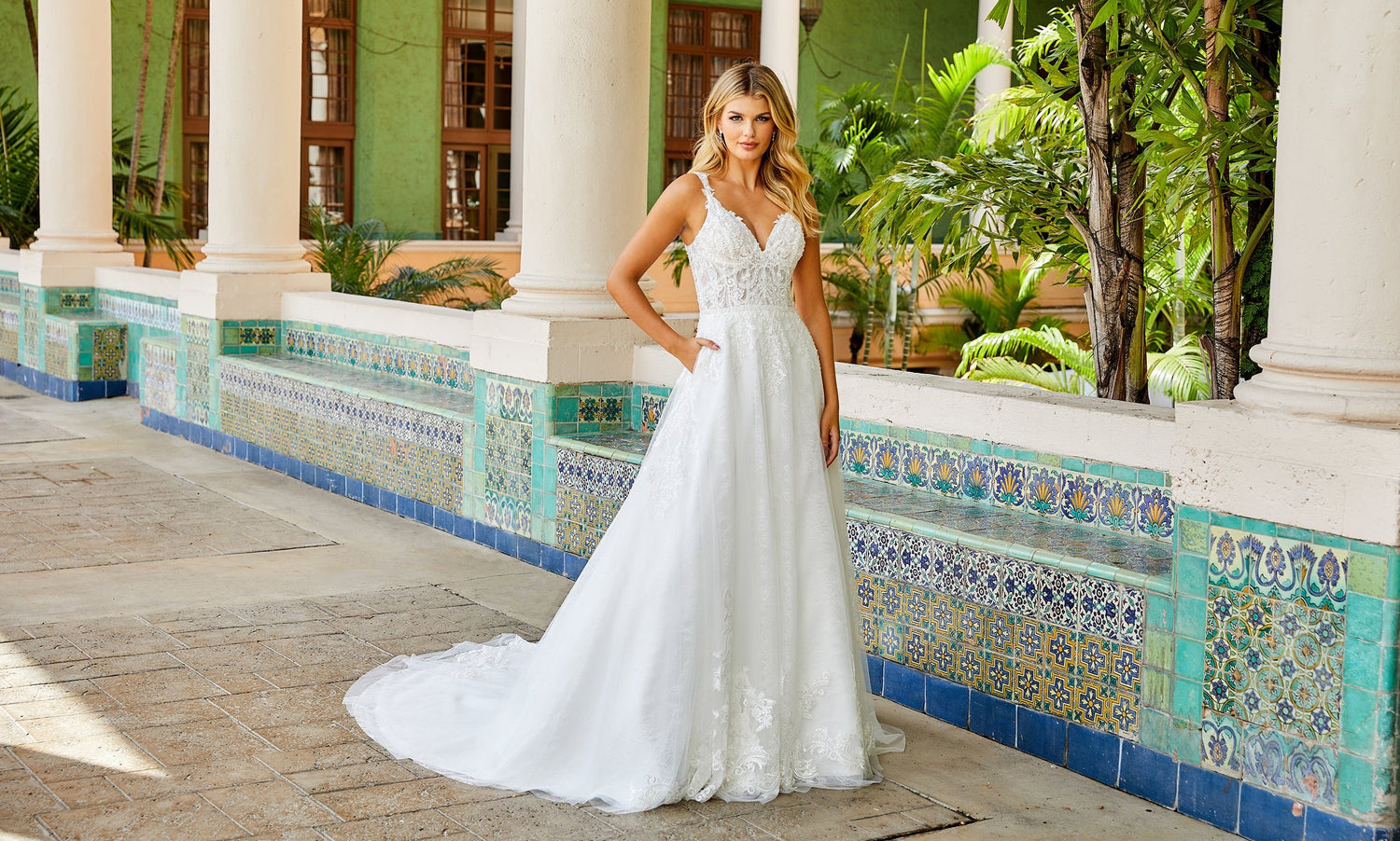 Blonde bride in a V-neck white lace A-line gown with pockets against a blue tiled wall