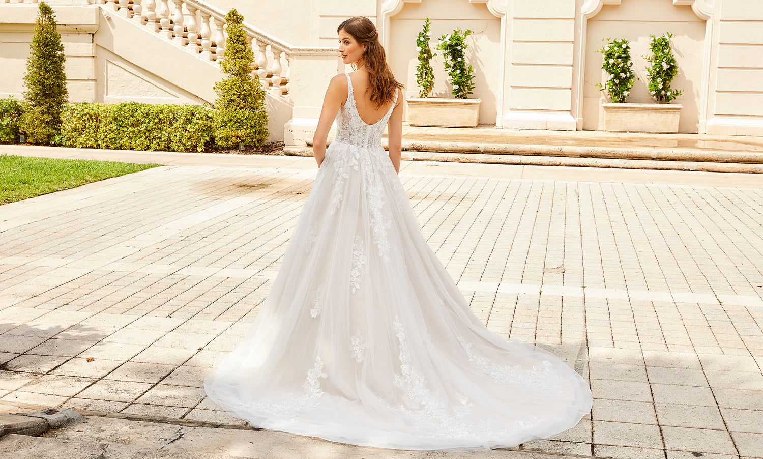 Back view of a bride in a scoop-back lace A-line gown with a long train on a paved courtyard
