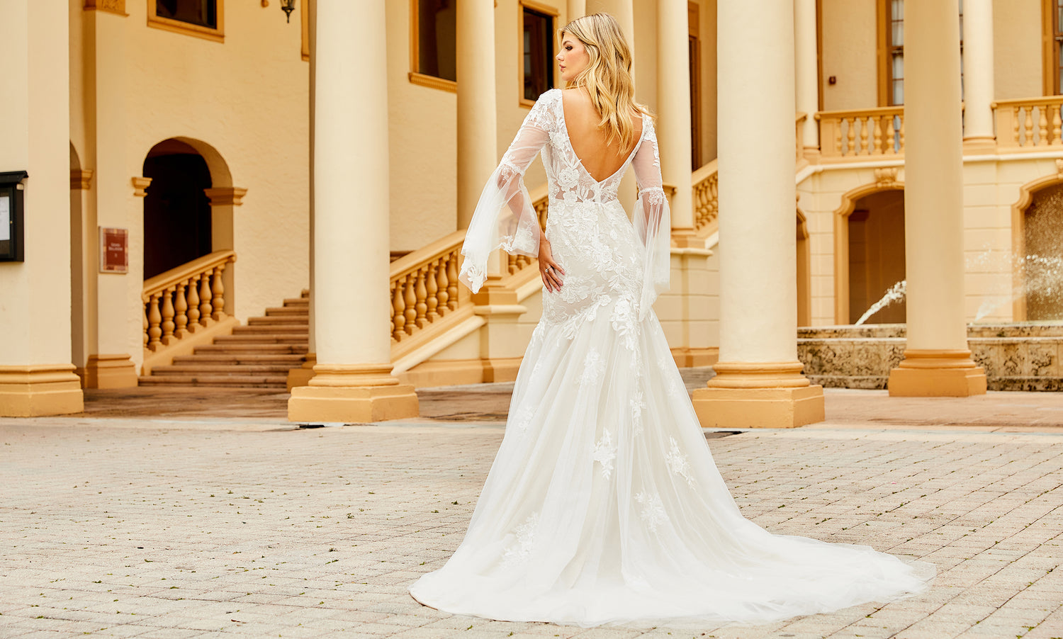 Back view of a bride in a deep V-back lace gown with sheer bell sleeves and a long train