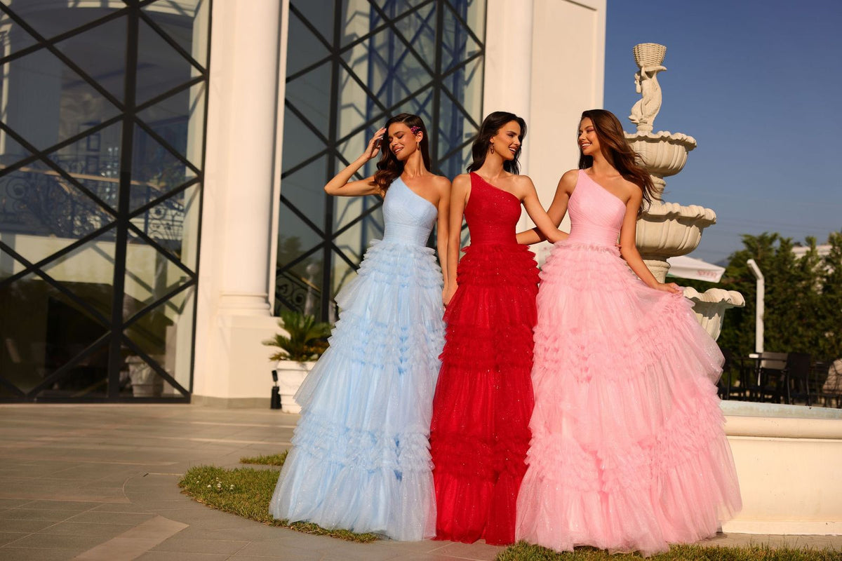 Three models in tiered ruffled tulle ball gowns, pale blue, red and pink, by a fountain
