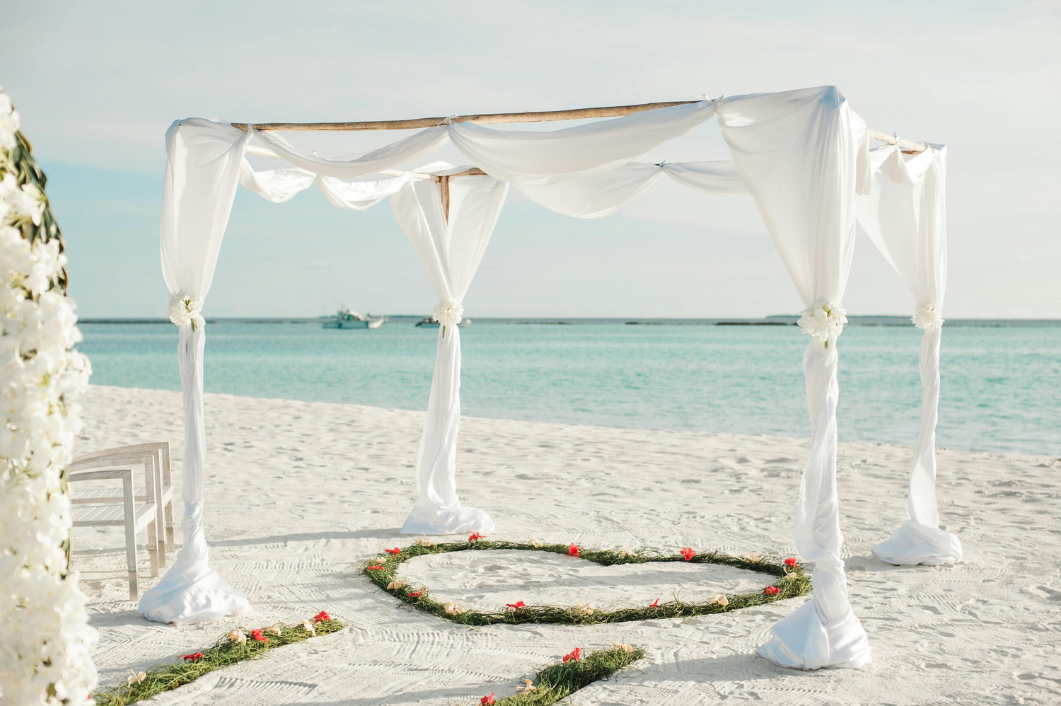 White draped ceremony canopy on a white sand beach beside turquoise water, ringed by greenery and pink petals