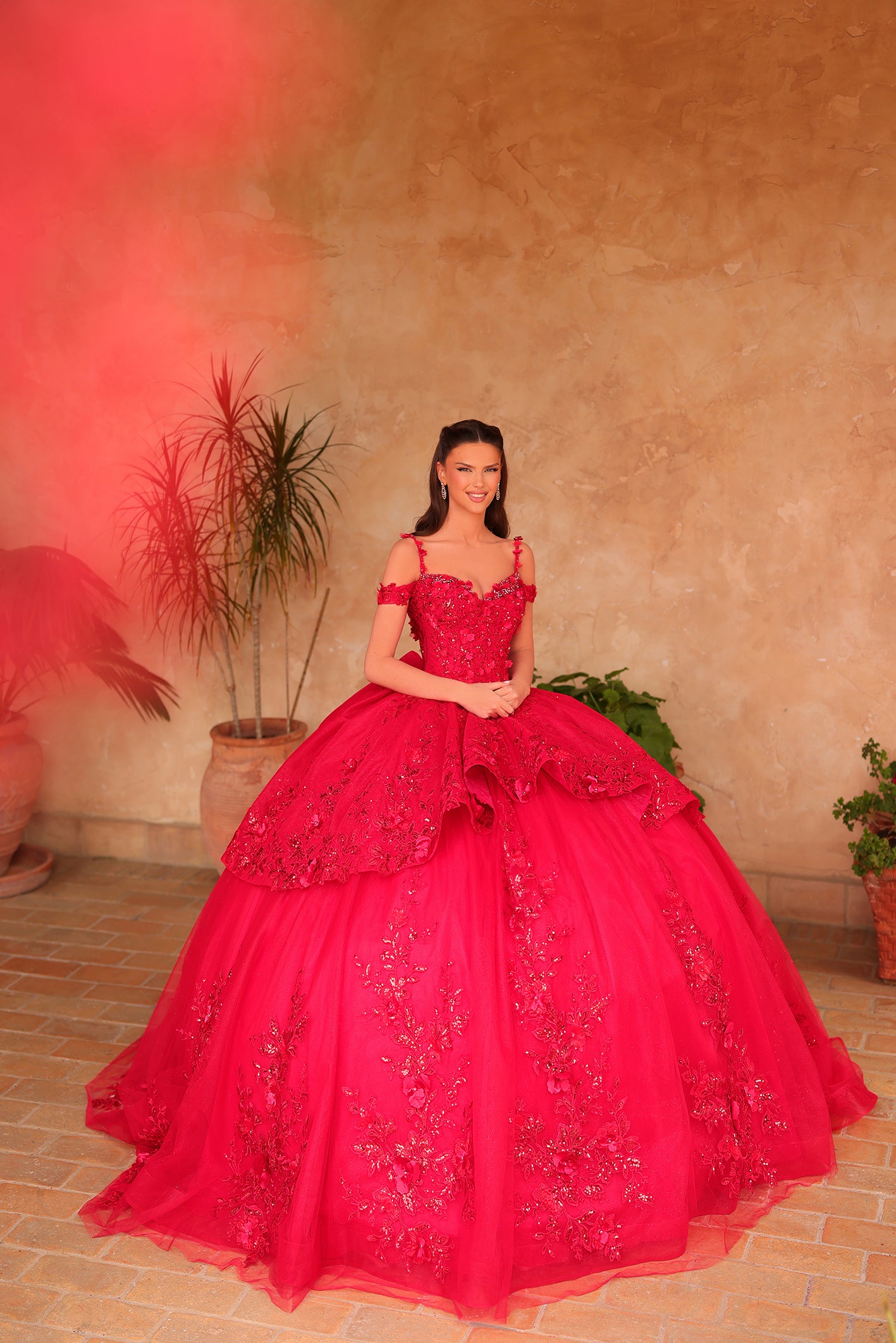 A woman wearing a voluminous red quinceañera gown stands in a terracotta-toned room with potted plants.