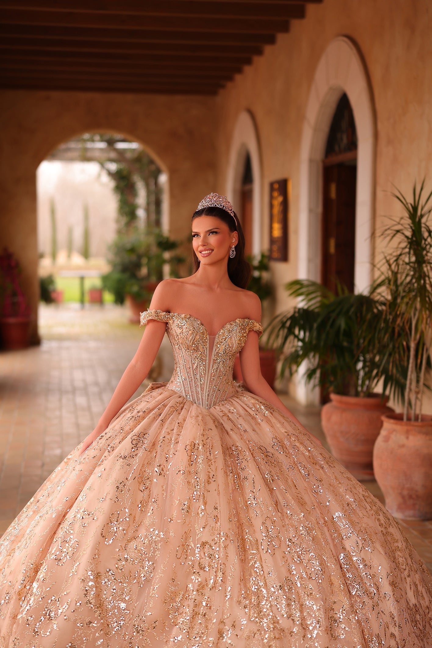 A glittering blush quinceañera ball gown with an off-shoulder bodice and sparkling embellishments in an arched courtyard.