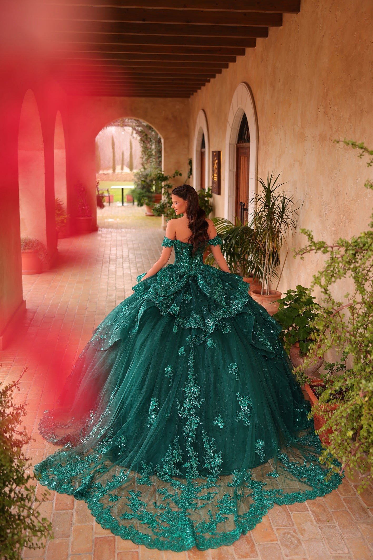 A woman wears an emerald green ballgown with intricate lace, standing in an arched courtyard with plants.