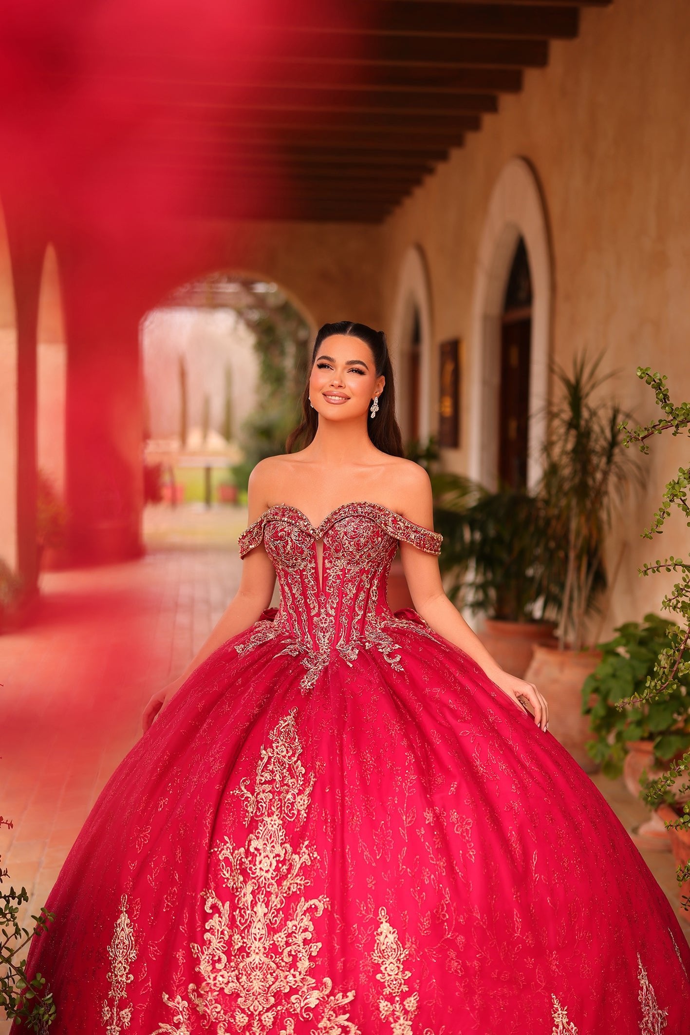 A woman wears an ornate red ball gown with silver embroidery, standing in an arched hallway with plants.