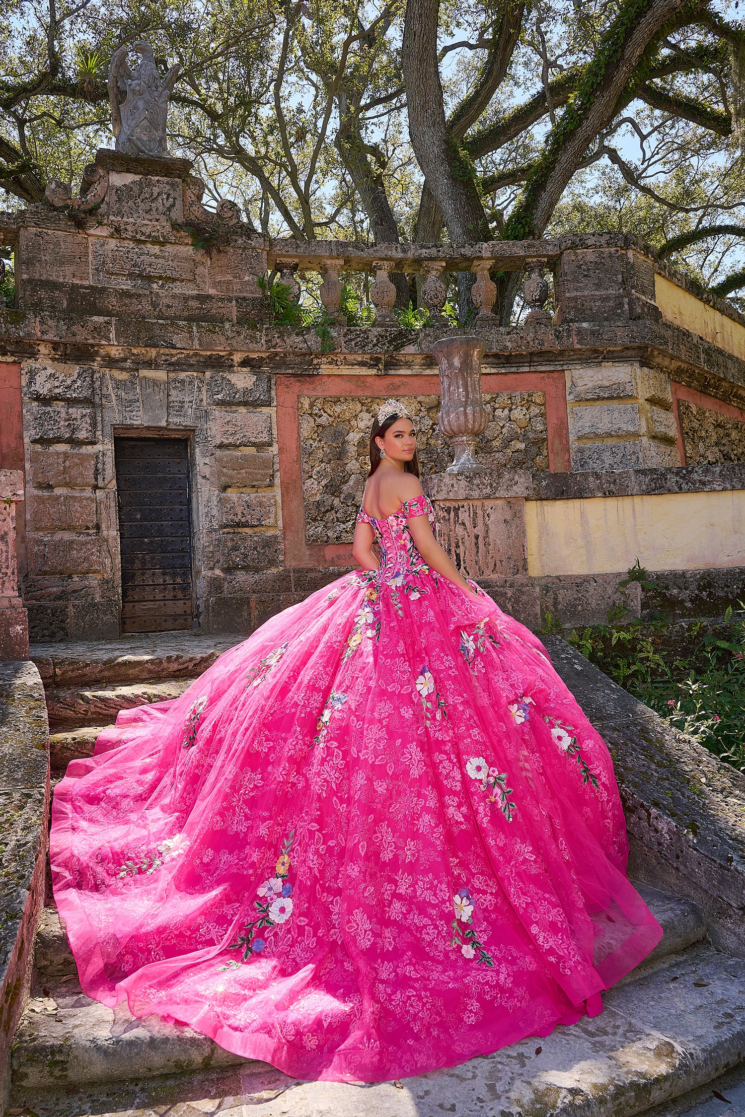 A woman in a vibrant pink floral quinceañera gown poses on stone steps with an ornate historic backdrop.