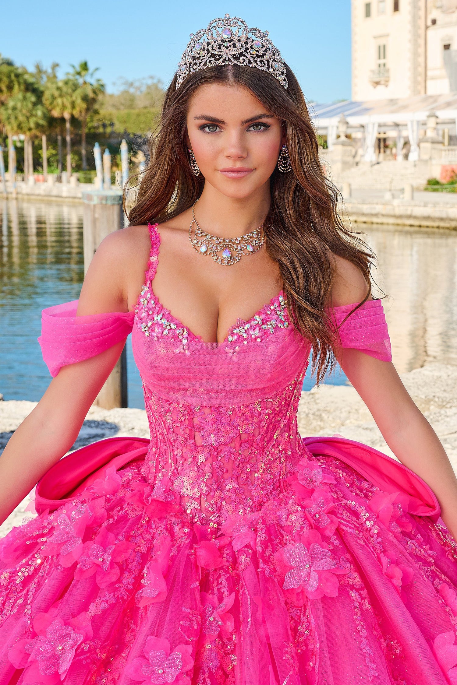 A woman in a vibrant pink quinceañera gown wears a sparkling tiara by a reflective water feature.