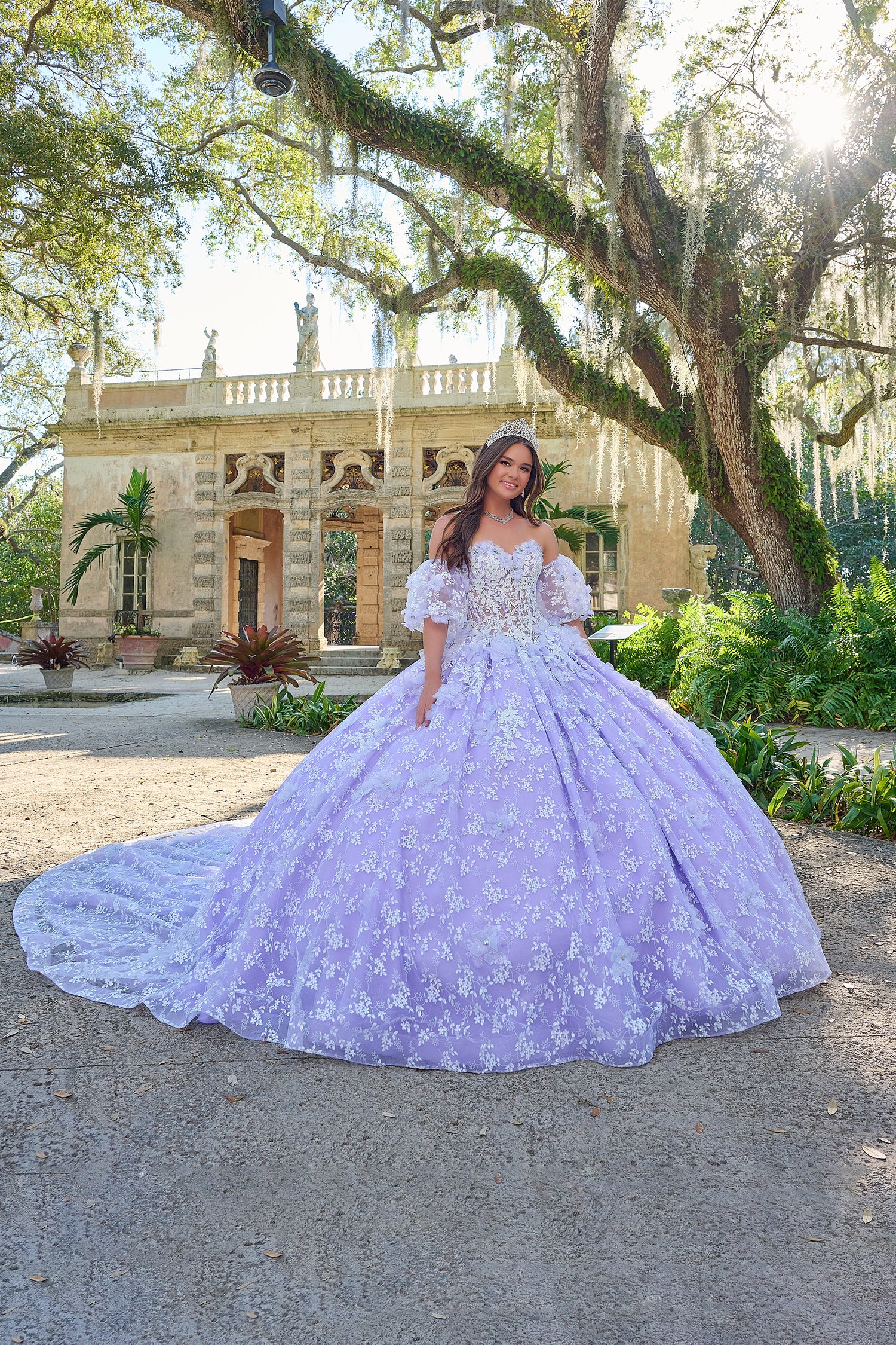 A young woman in a lilac quinceañera gown poses in a lush garden with a historic stone building behind her.