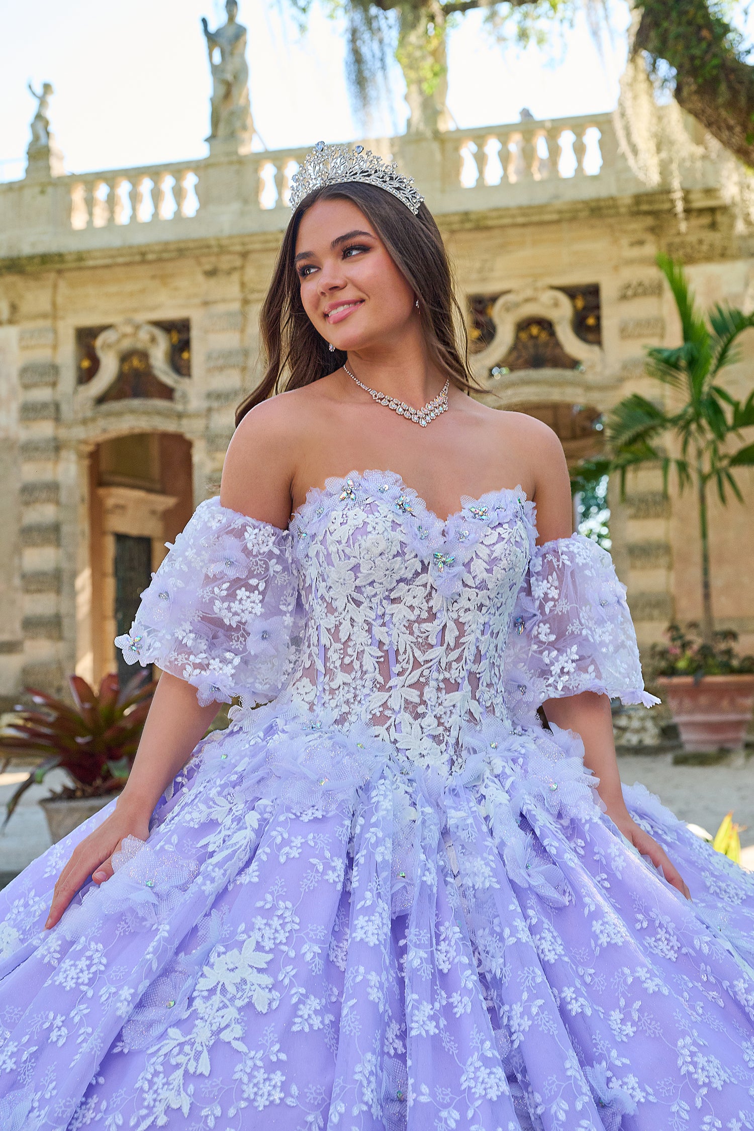 A young woman in a lavender quinceañera gown with floral lace and a tiara poses near a classical building.