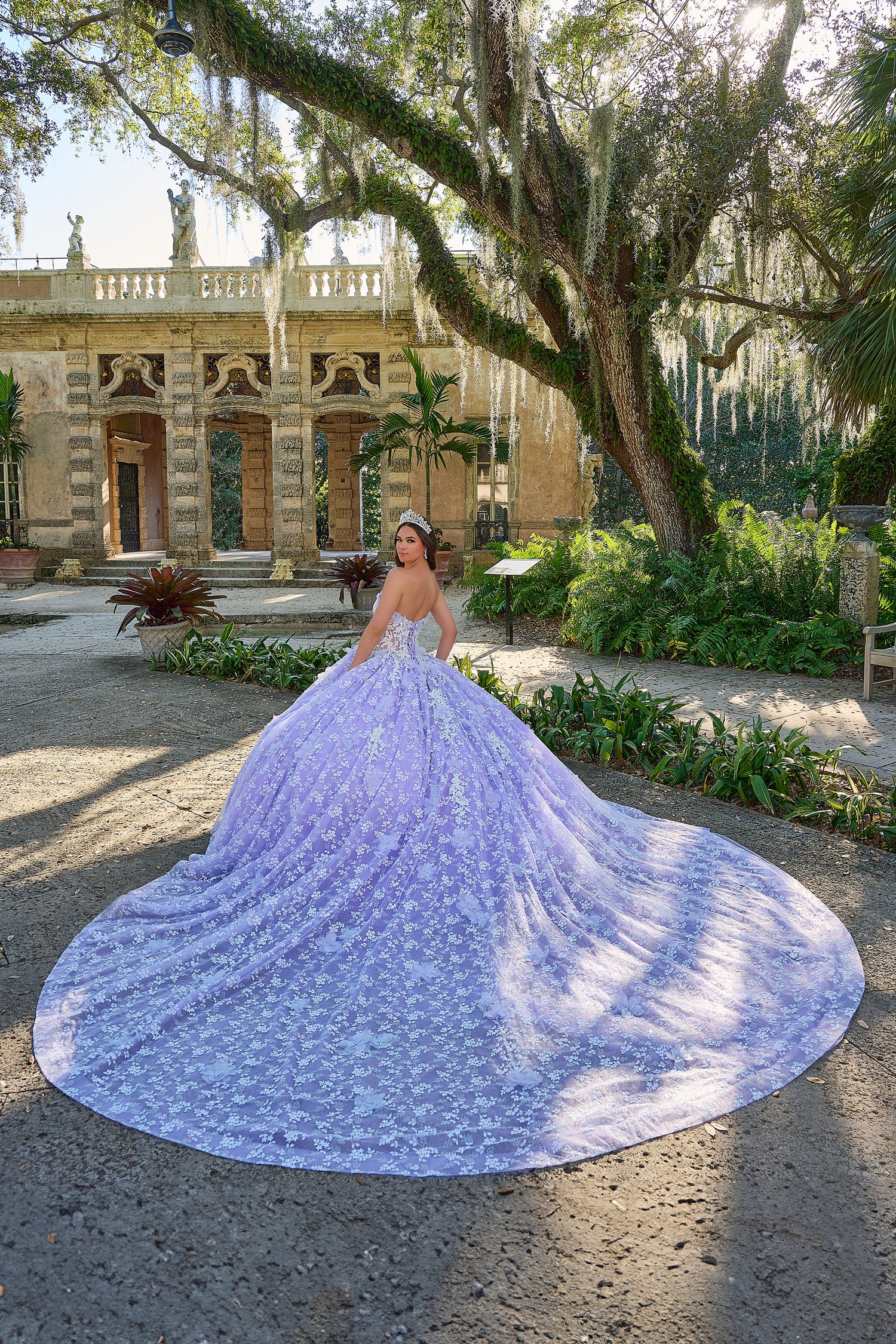 A lavender quinceañera gown spreads dramatically in a lush garden with an ornate historic building behind.