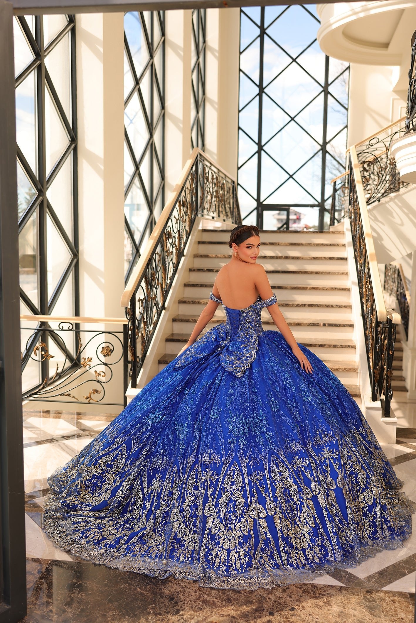 A woman in a royal blue ballgown with gold embellishments poses on an elegant staircase with ornate railings.