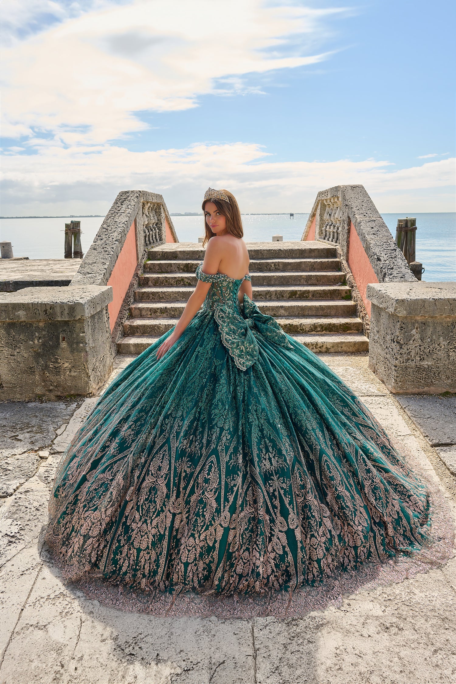 A woman in a teal embellished ballgown poses on stone steps by the ocean, wearing a tiara.