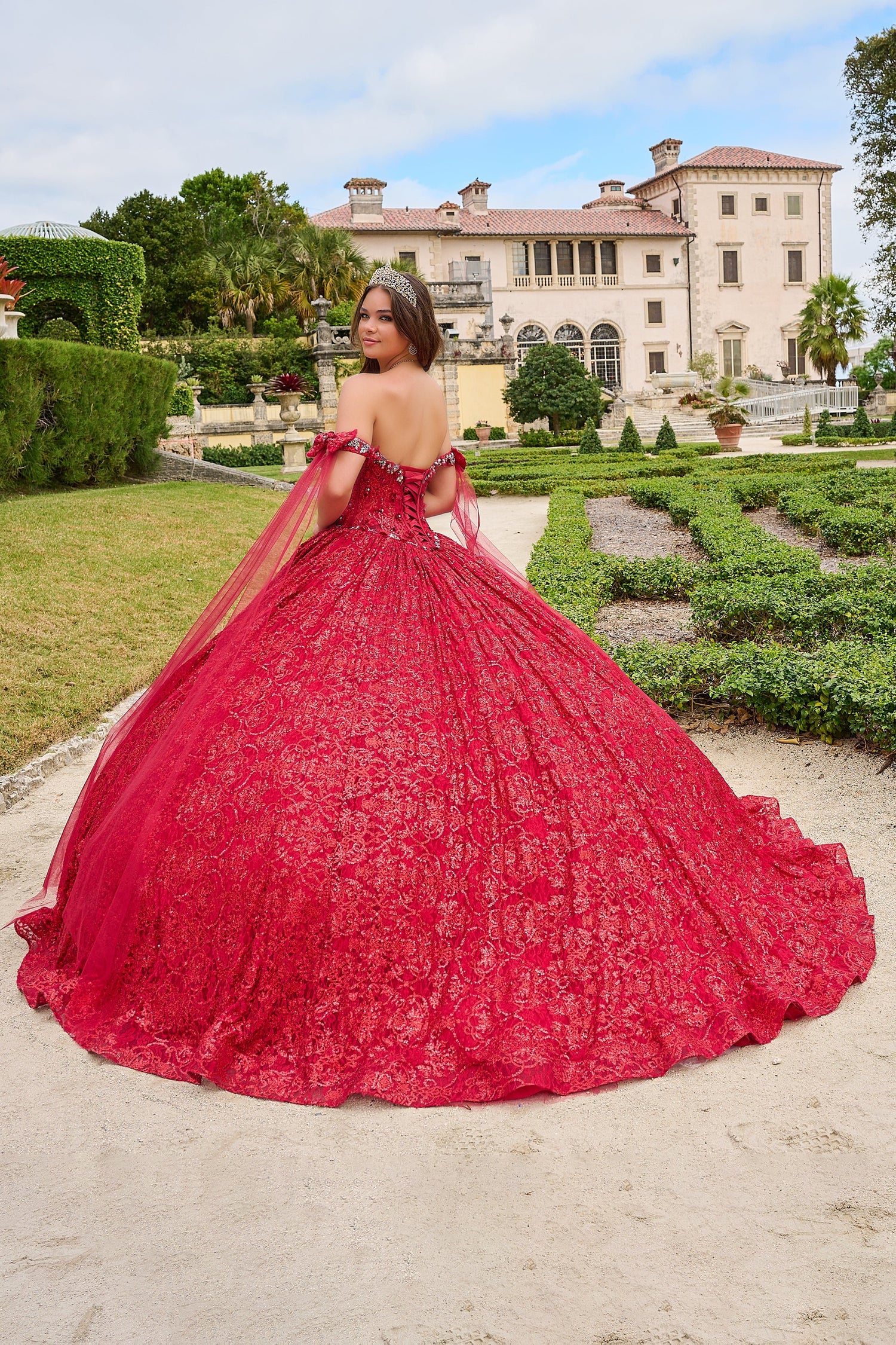 A woman in a vibrant red ball gown and tiara poses in a manicured garden with an elegant mansion backdrop.