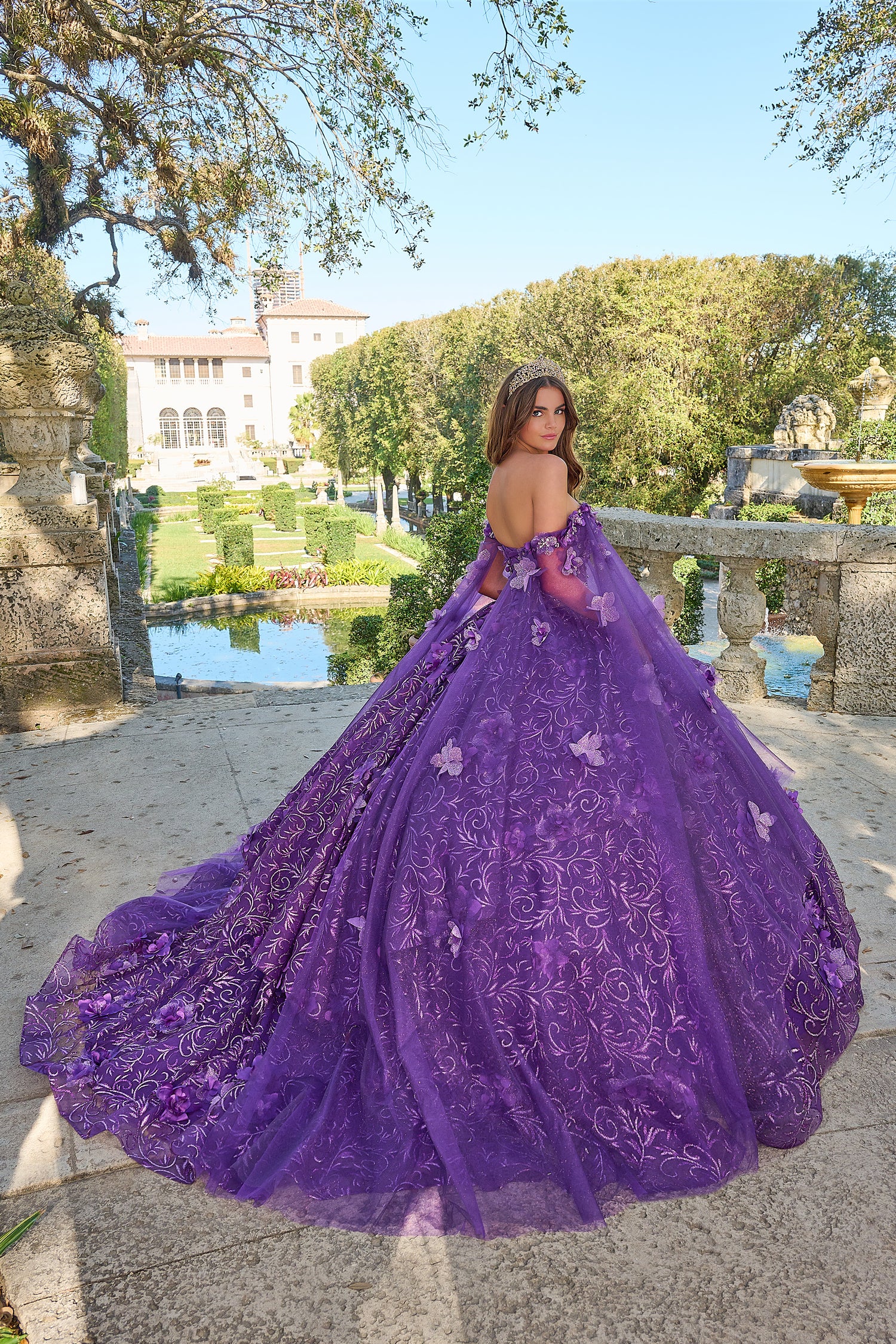 A woman in a voluminous purple off-shoulder ball gown poses in a garden with a historic mansion in the background.