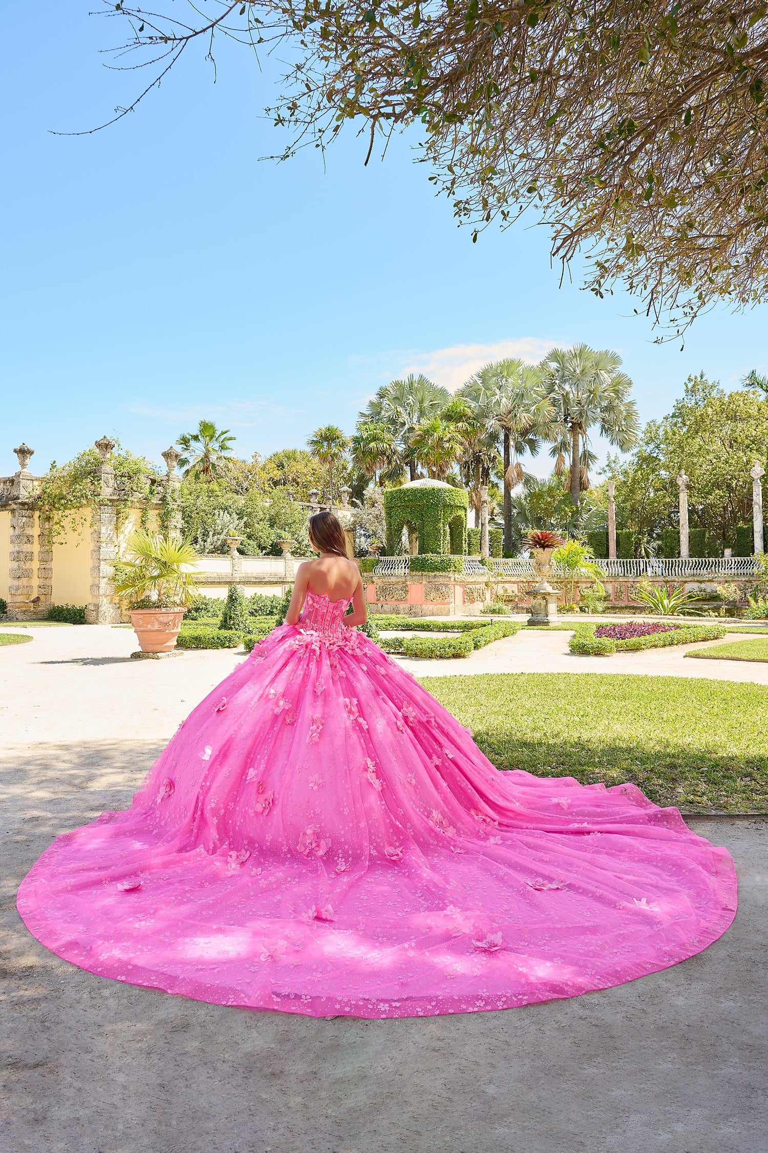 A person in a voluminous pink floral gown stands in a manicured garden with palm trees and stone walls.