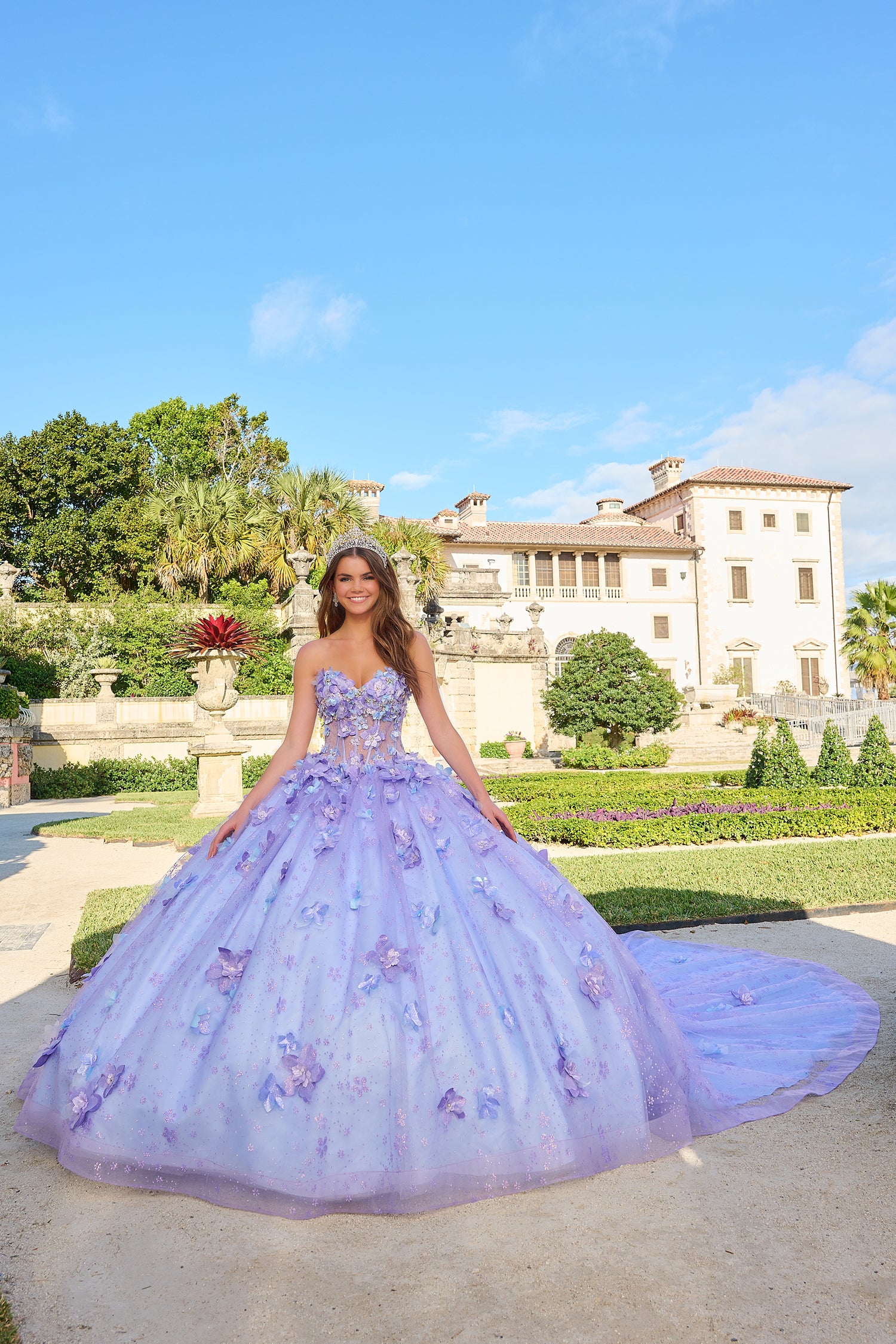 A young woman in a lavender butterfly-adorned ball gown poses in a manicured garden with a grand villa behind her.