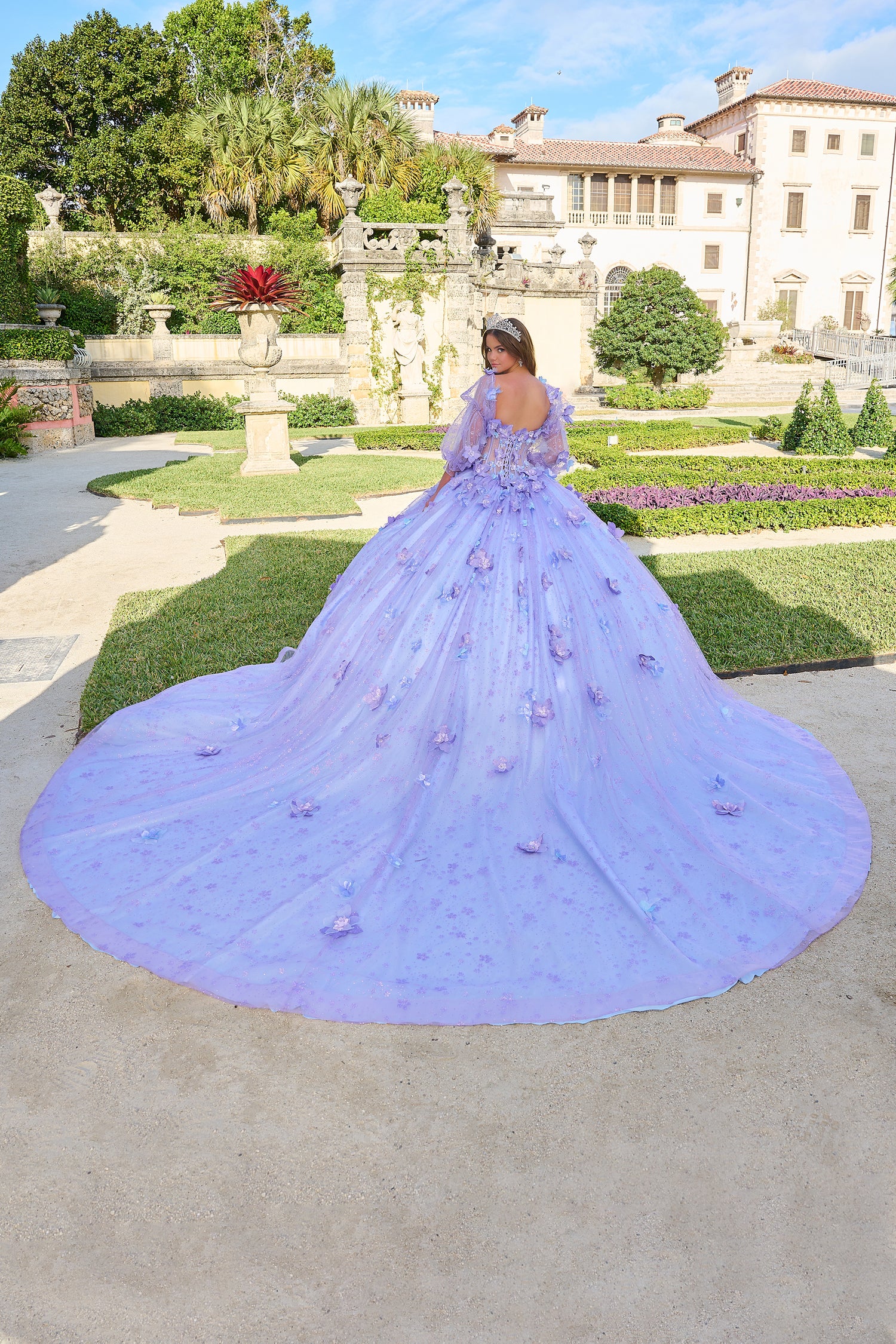 A lavender ball gown with 3D floral details stands in an elegant garden with a historic mansion backdrop.