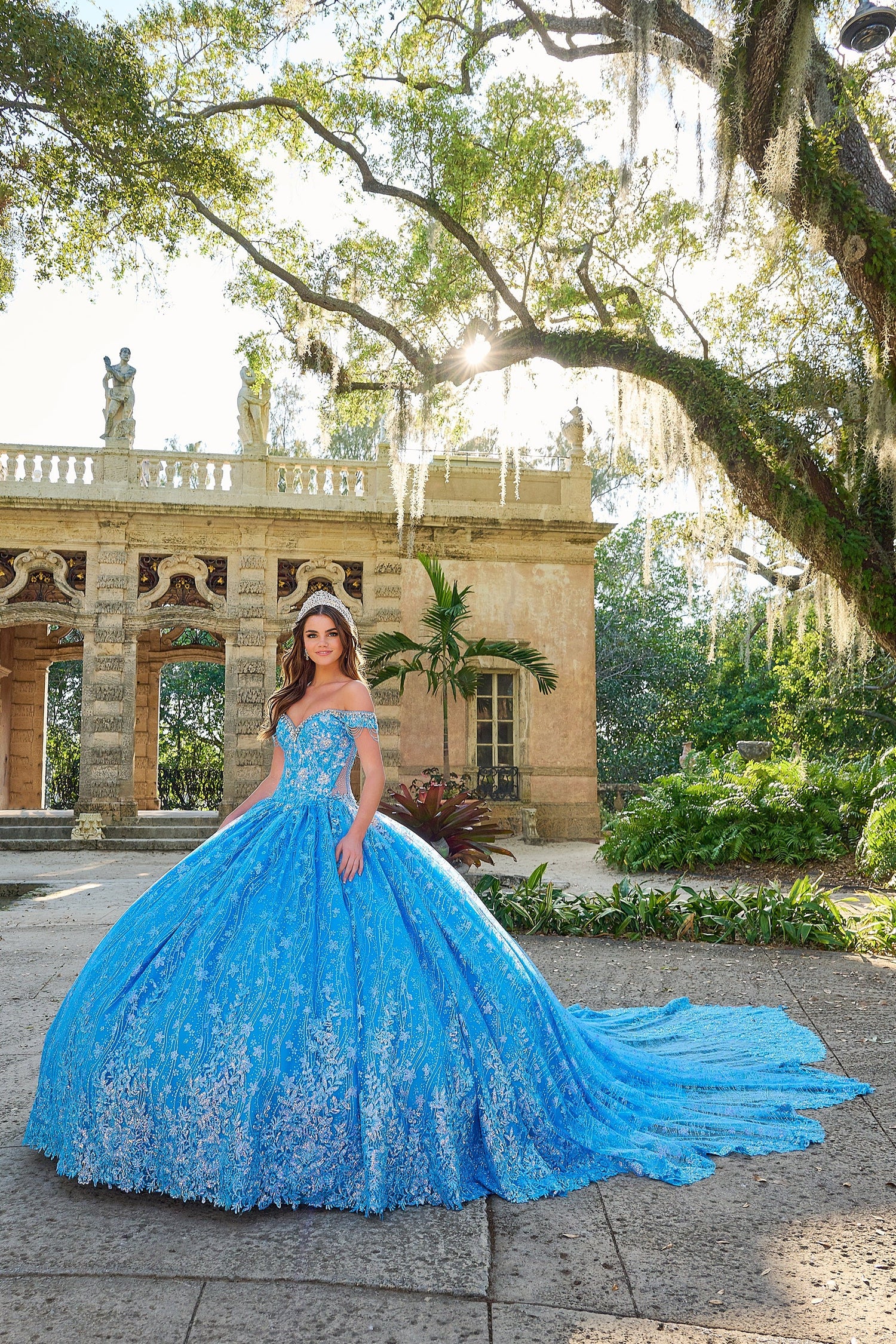 A young woman in a turquoise lace ball gown poses in a garden with ornate architecture and trees.