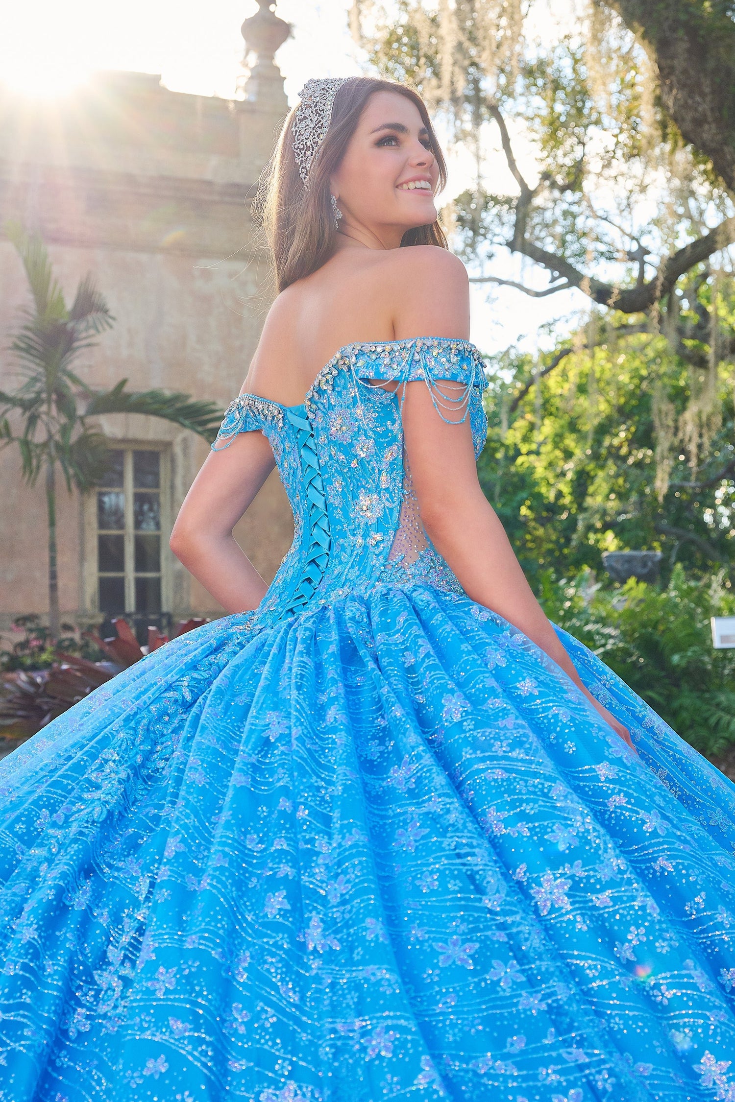 A woman in a sparkly blue off-shoulder ball gown stands in a garden with ornate architecture behind her.