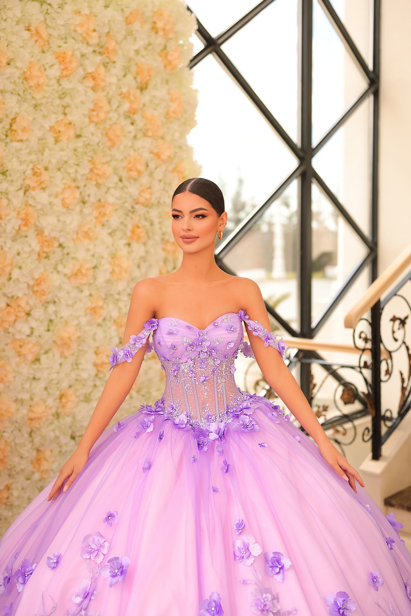 A woman wears a lavender ball gown with 3D floral details and a sheer corset bodice against a flower wall.