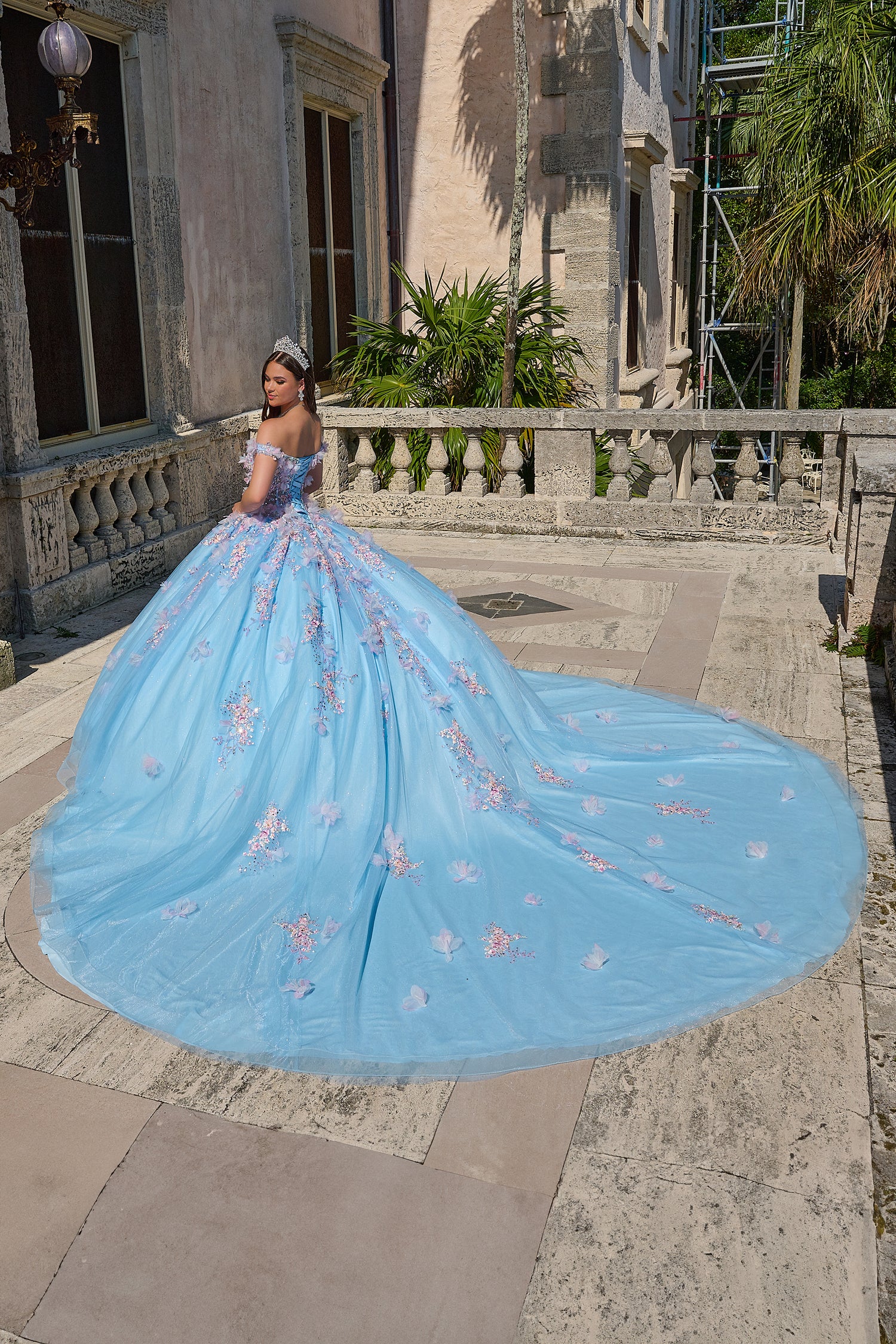 A blue off-shoulder ball gown with pink floral embroidery and a long train, posed on a stone terrace.