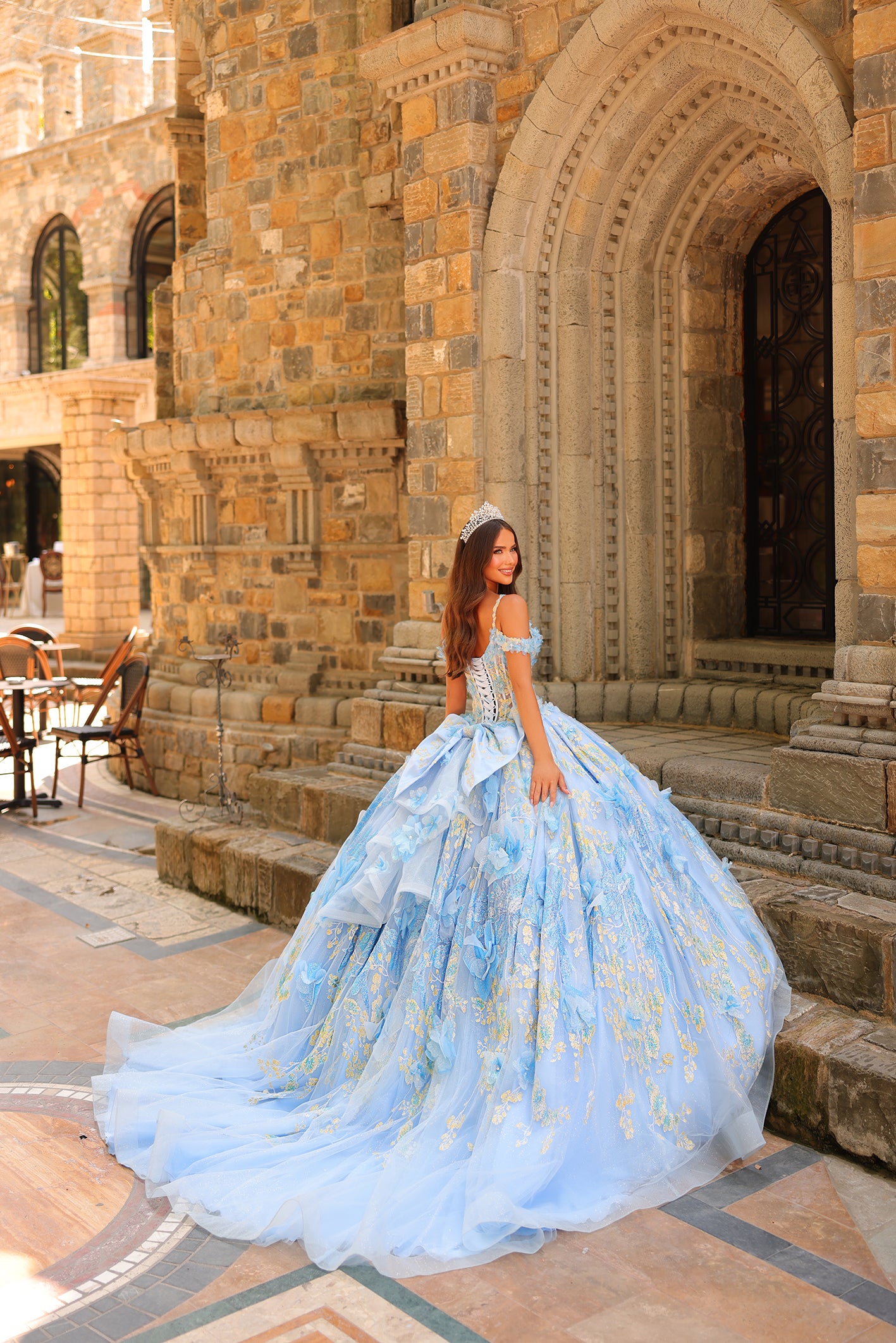 A woman in a voluminous light blue ball gown and tiara poses before an ornate stone building.