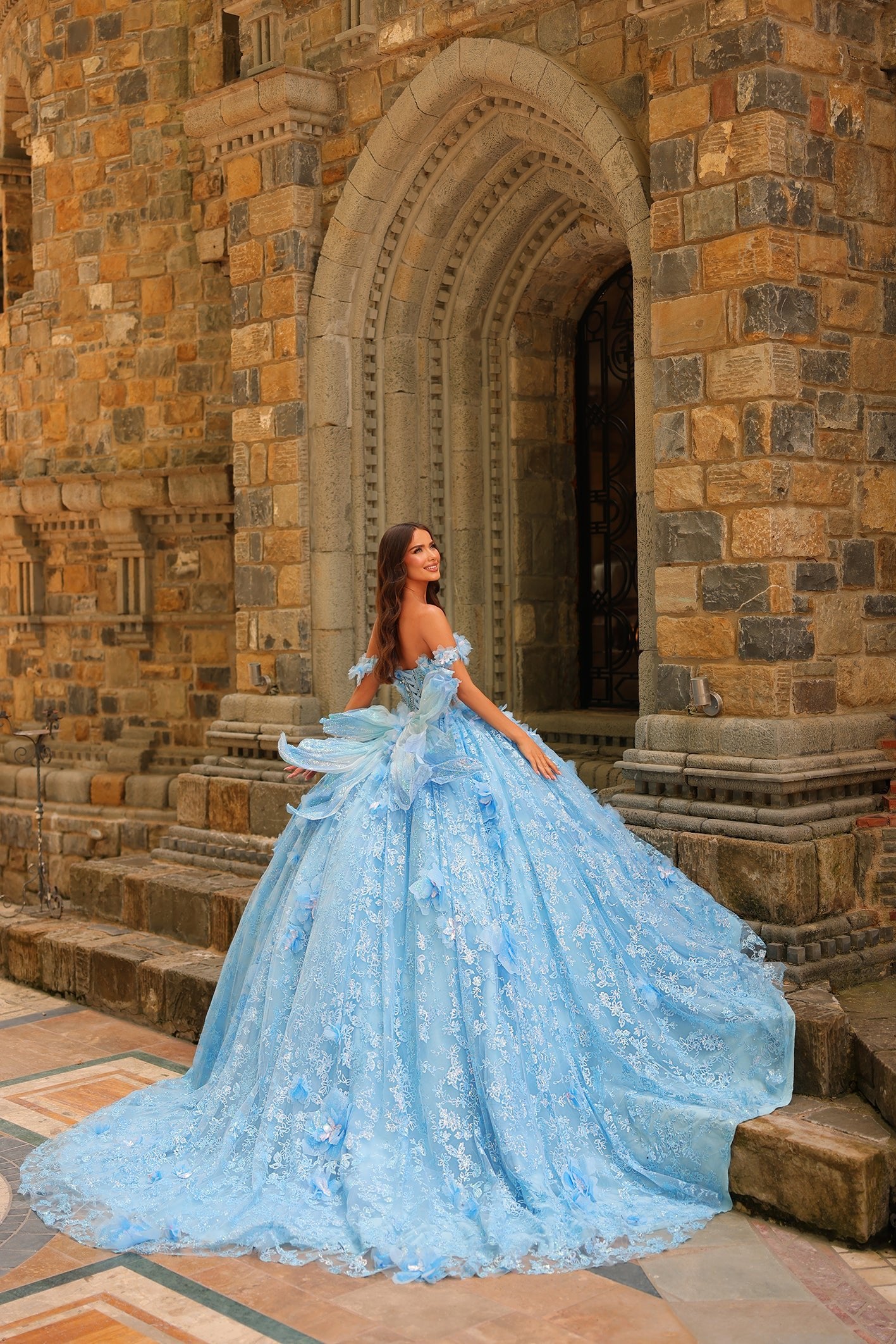 A woman in a voluminous blue lace ballgown poses on stone steps near an ornate medieval archway.