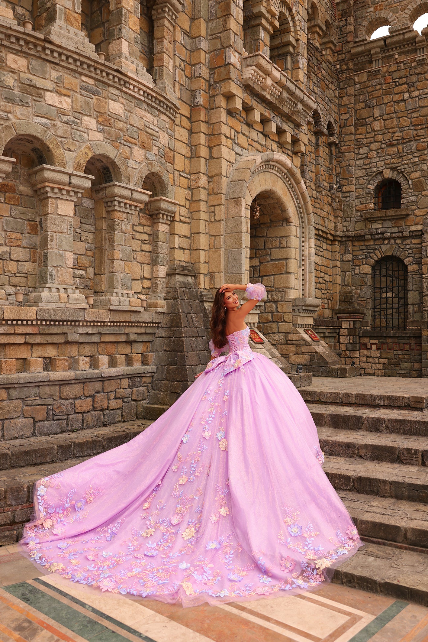 A woman in a lavender ball gown stands on stone steps of a medieval castle, posing with a sweeping floral dress train.