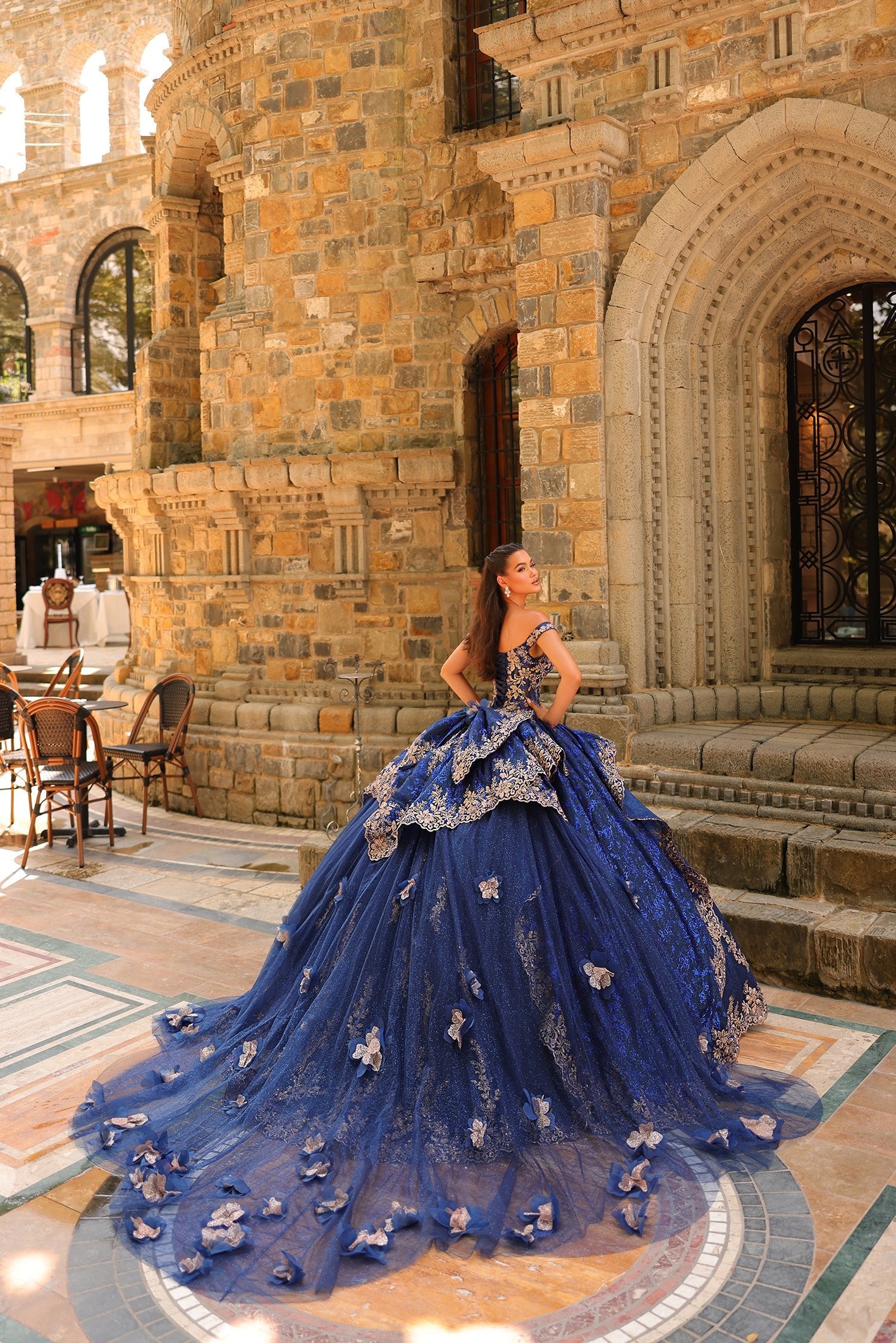 A young woman in an elaborate royal blue ball gown poses dramatically in a stone architectural setting.