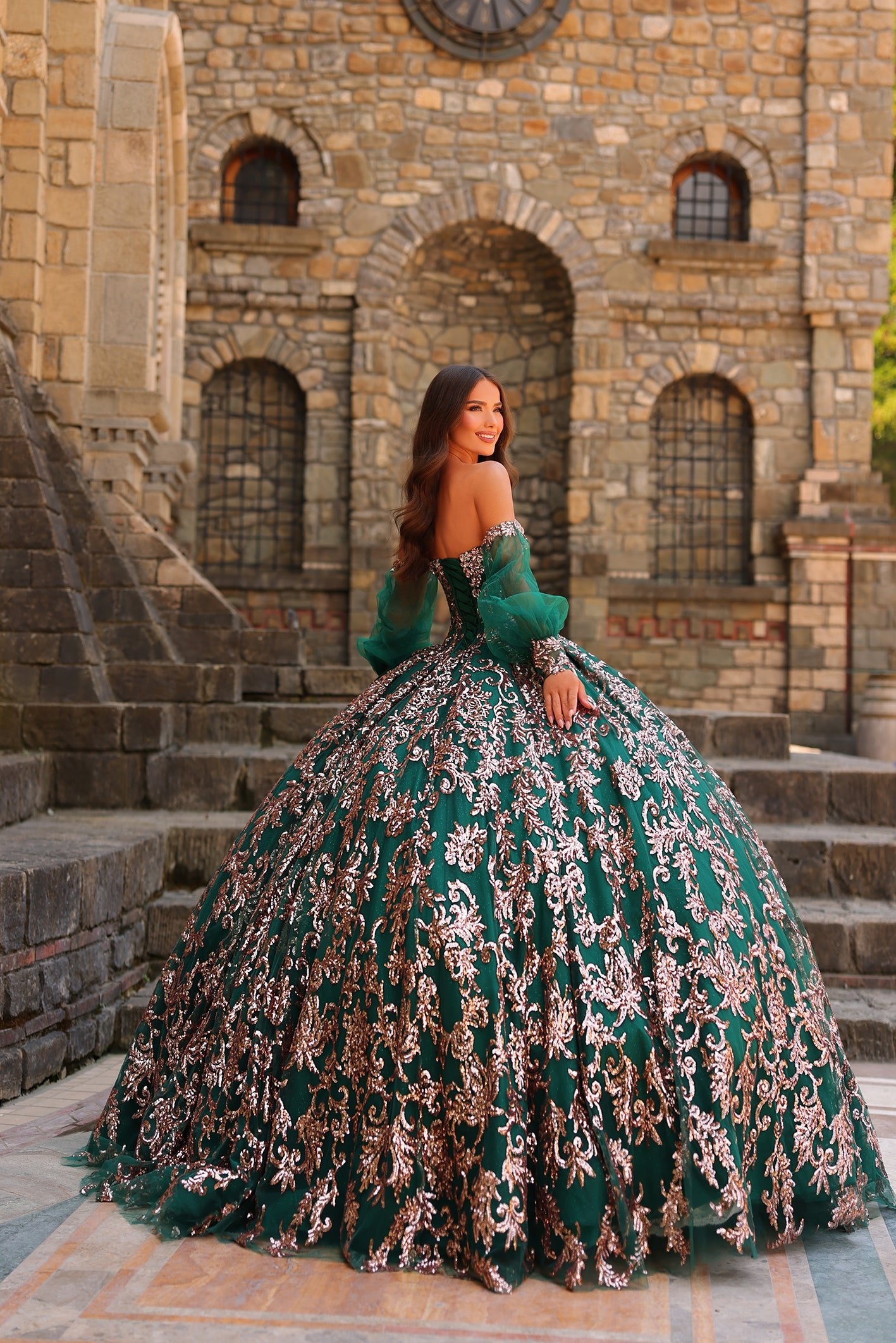 A woman in an emerald green sequined ballgown stands on stone steps before a medieval castle wall.