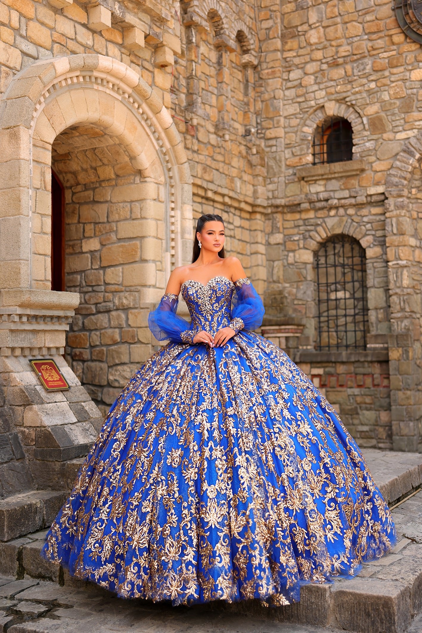 A woman wears a royal blue ball gown with gold embroidery, standing in front of a stone castle wall.