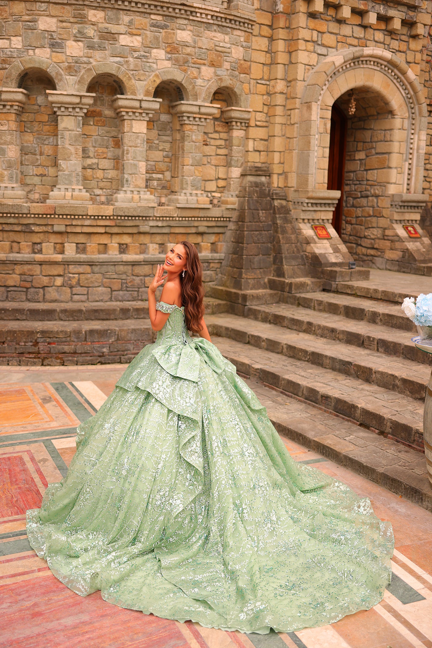 A woman in a sparkly sage green ballgown poses dramatically on stone steps in front of an ornate stone building.
