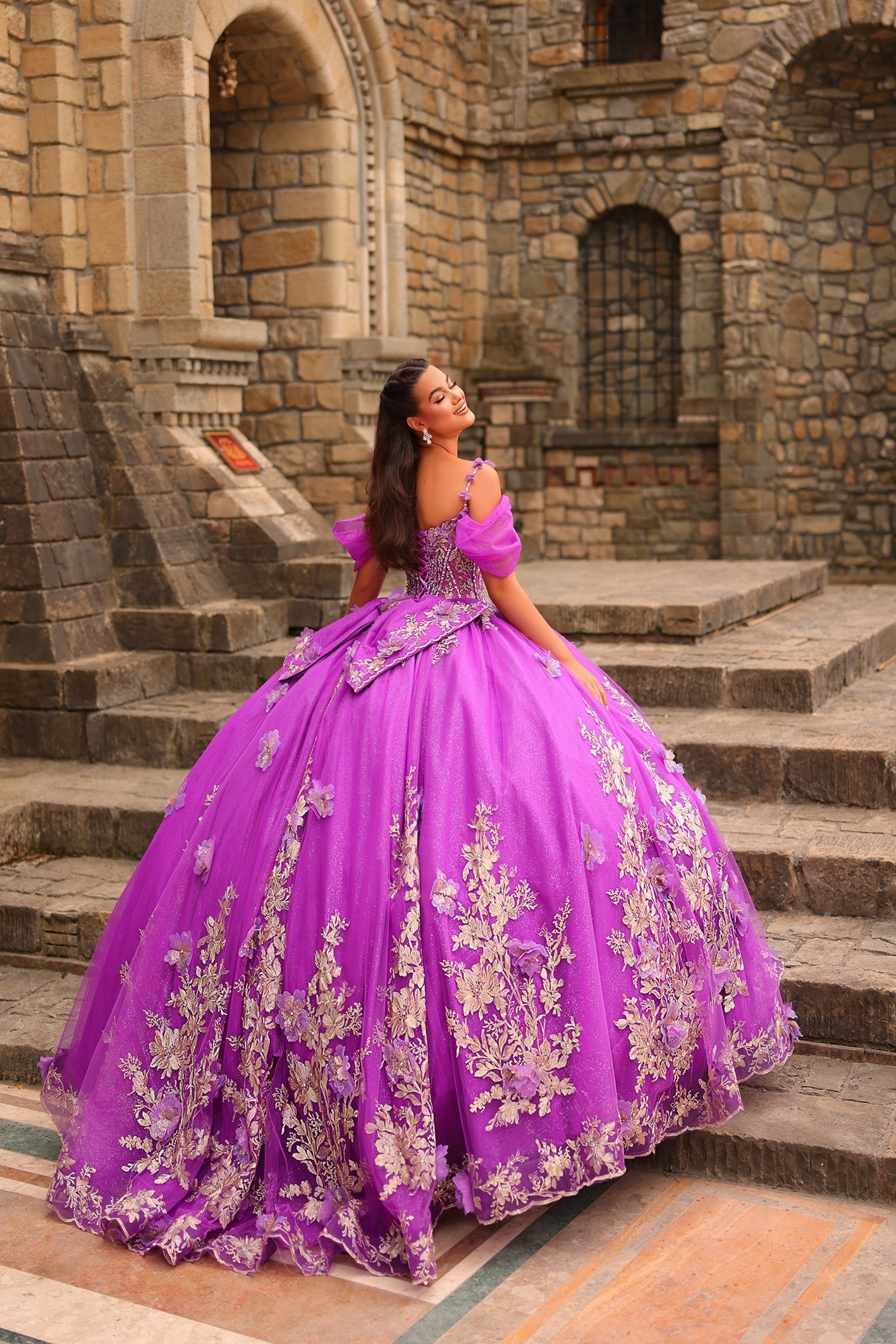 A woman in a vibrant purple ball gown with floral embroidery poses on stone steps near an ancient stone building.