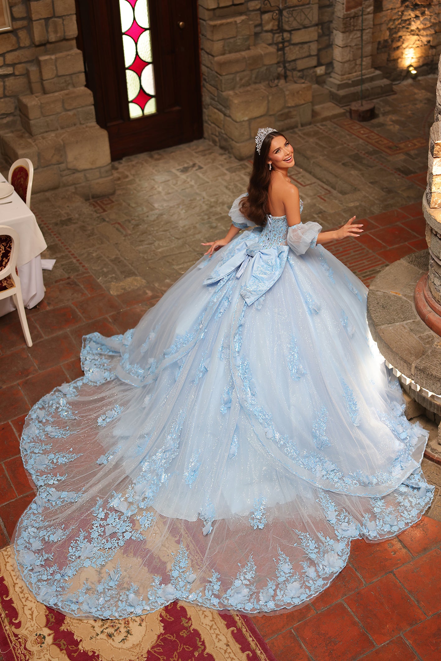 A blue ballgown with lace details and a long train, worn by a woman posing in a stone-walled room.