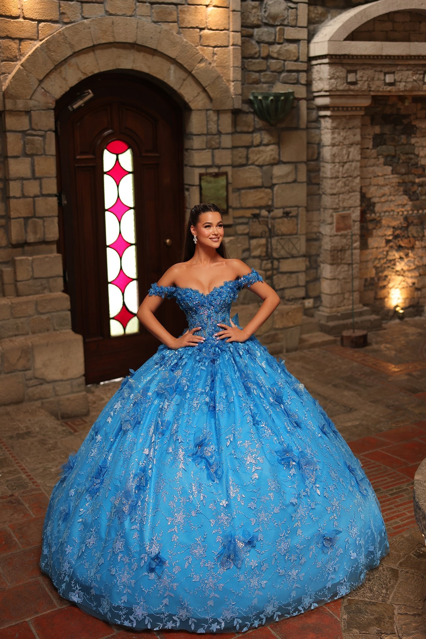 A woman wears a turquoise off-shoulder ball gown with intricate floral embroidery in a stone castle setting.