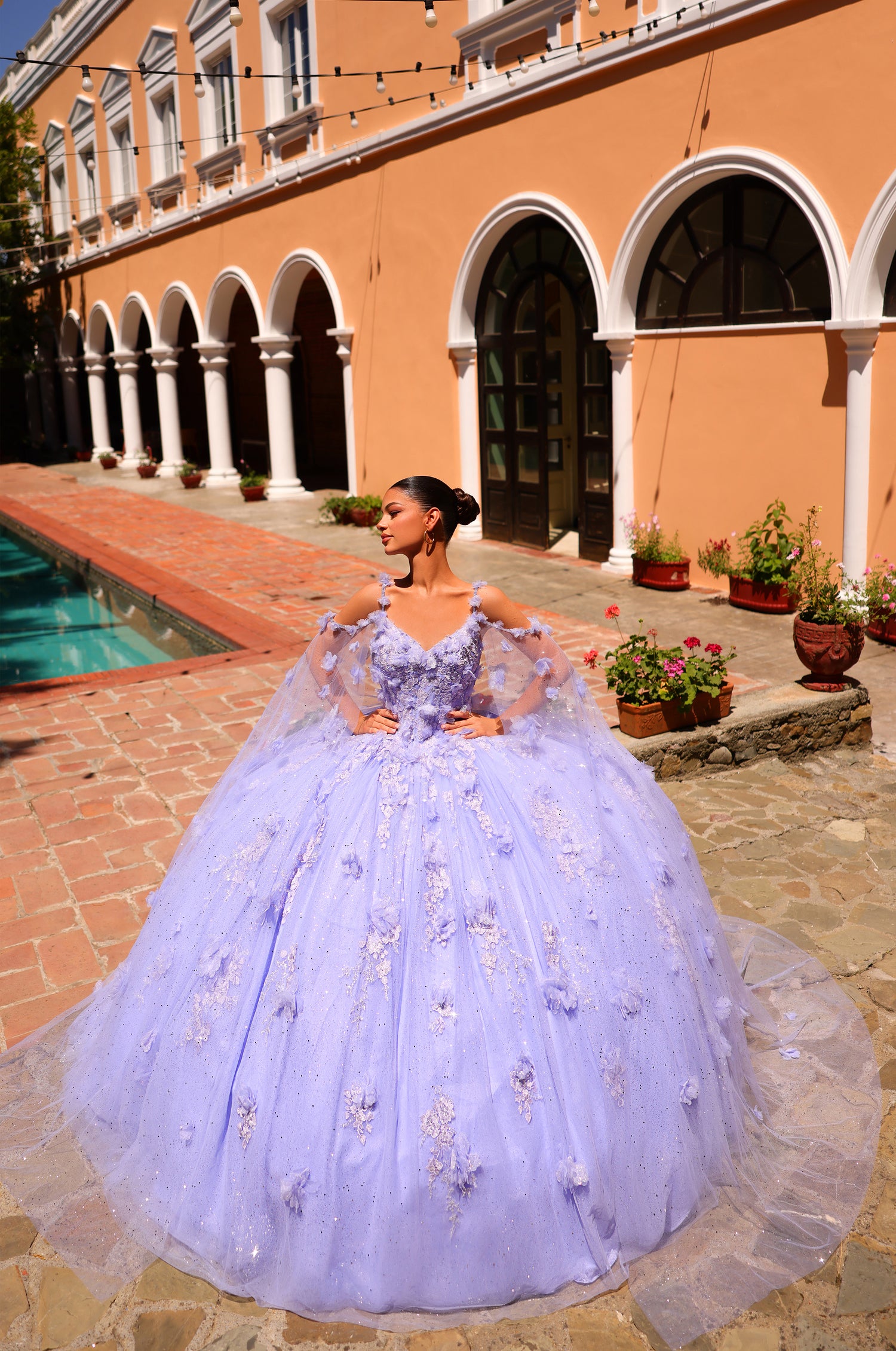 A lavender ball gown with 3D floral appliqués stands near a terracotta-colored building with arched windows.