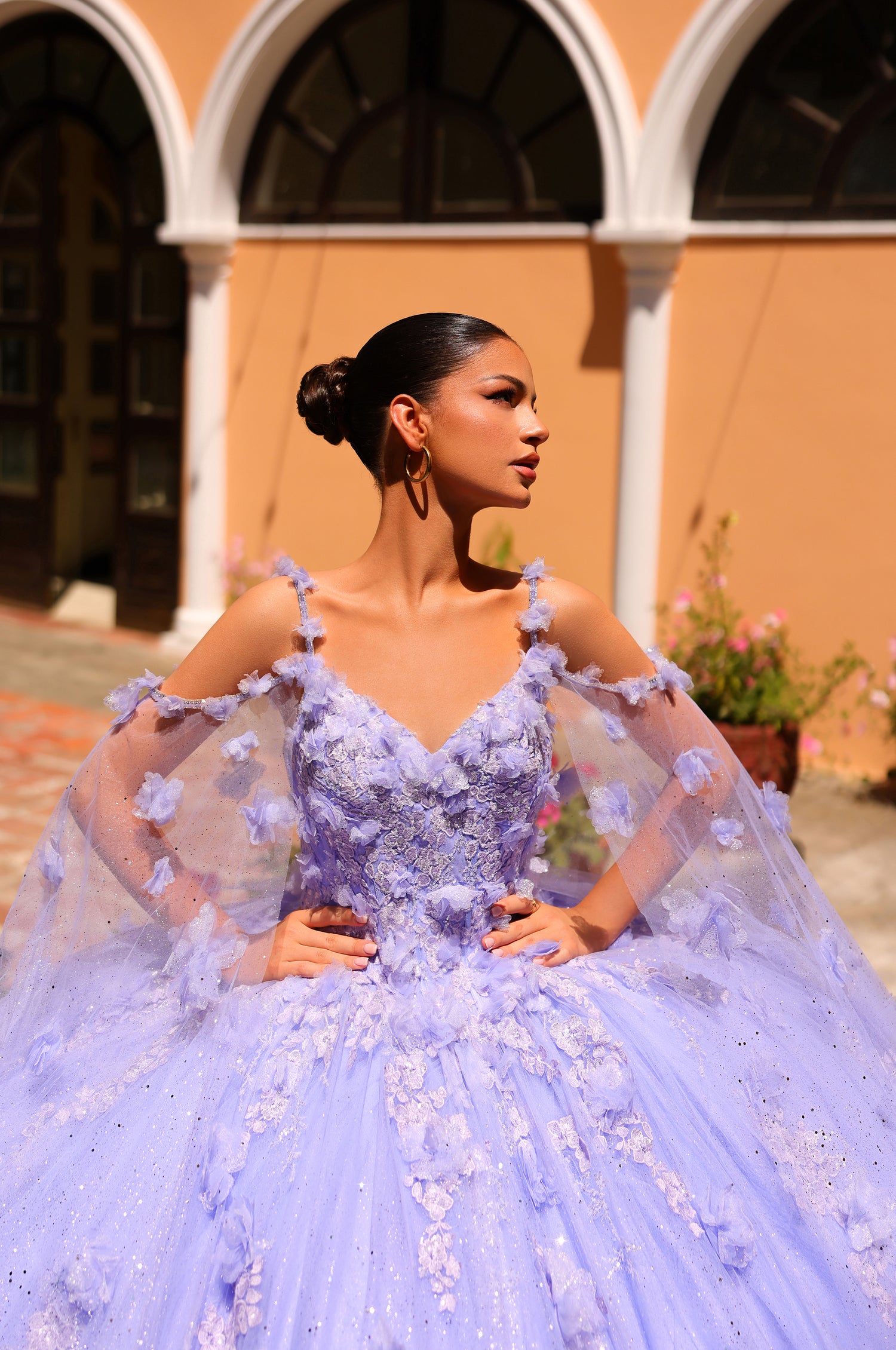 A woman in a lavender ball gown with 3D floral details poses elegantly against an arched architectural backdrop.