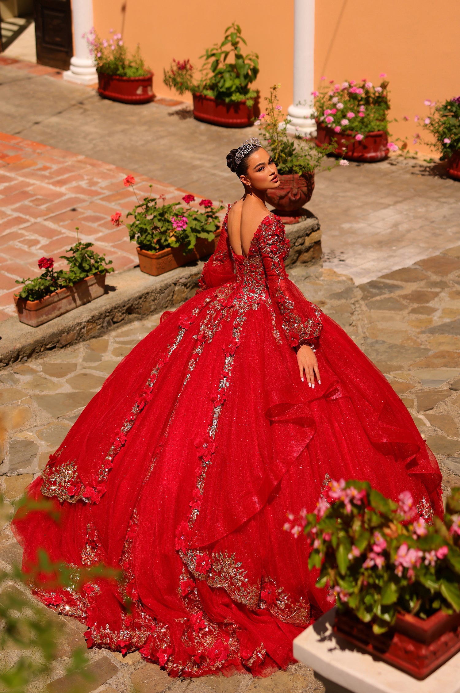 A woman in a vibrant red quinceañera gown with silver embroidery poses among flower-filled terracotta pots.