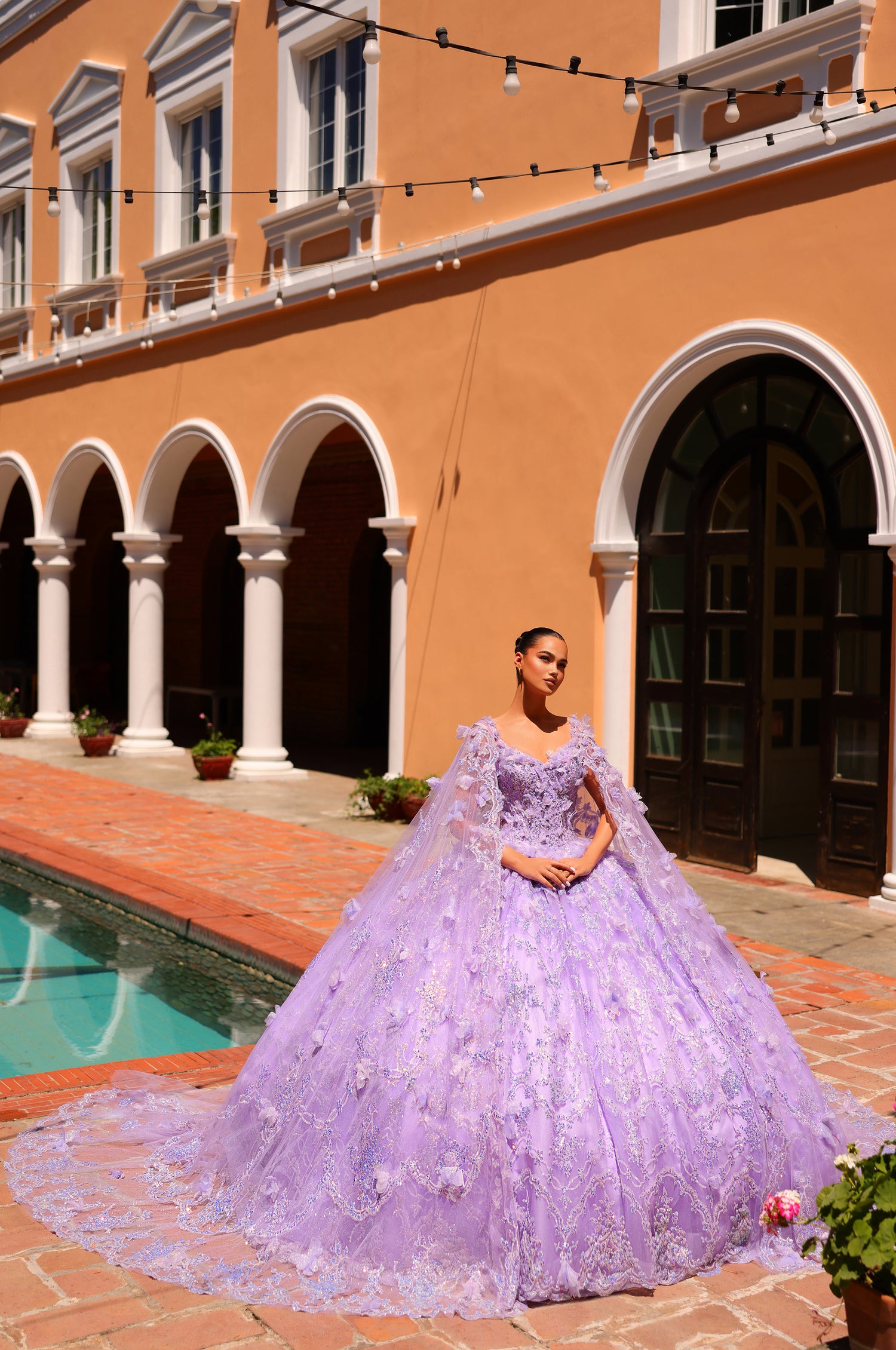 A woman in a lavender ball gown with 3D floral details poses by a Mediterranean-style building with arched windows.